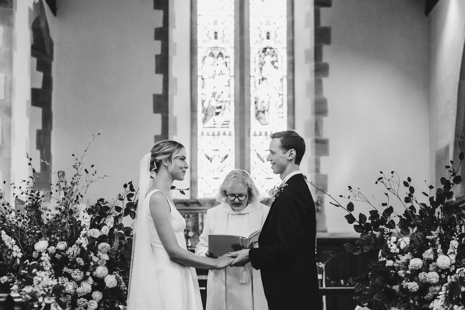 Black and white photo of wedding ceremony at altar with floral arrangements and stained glass windows.