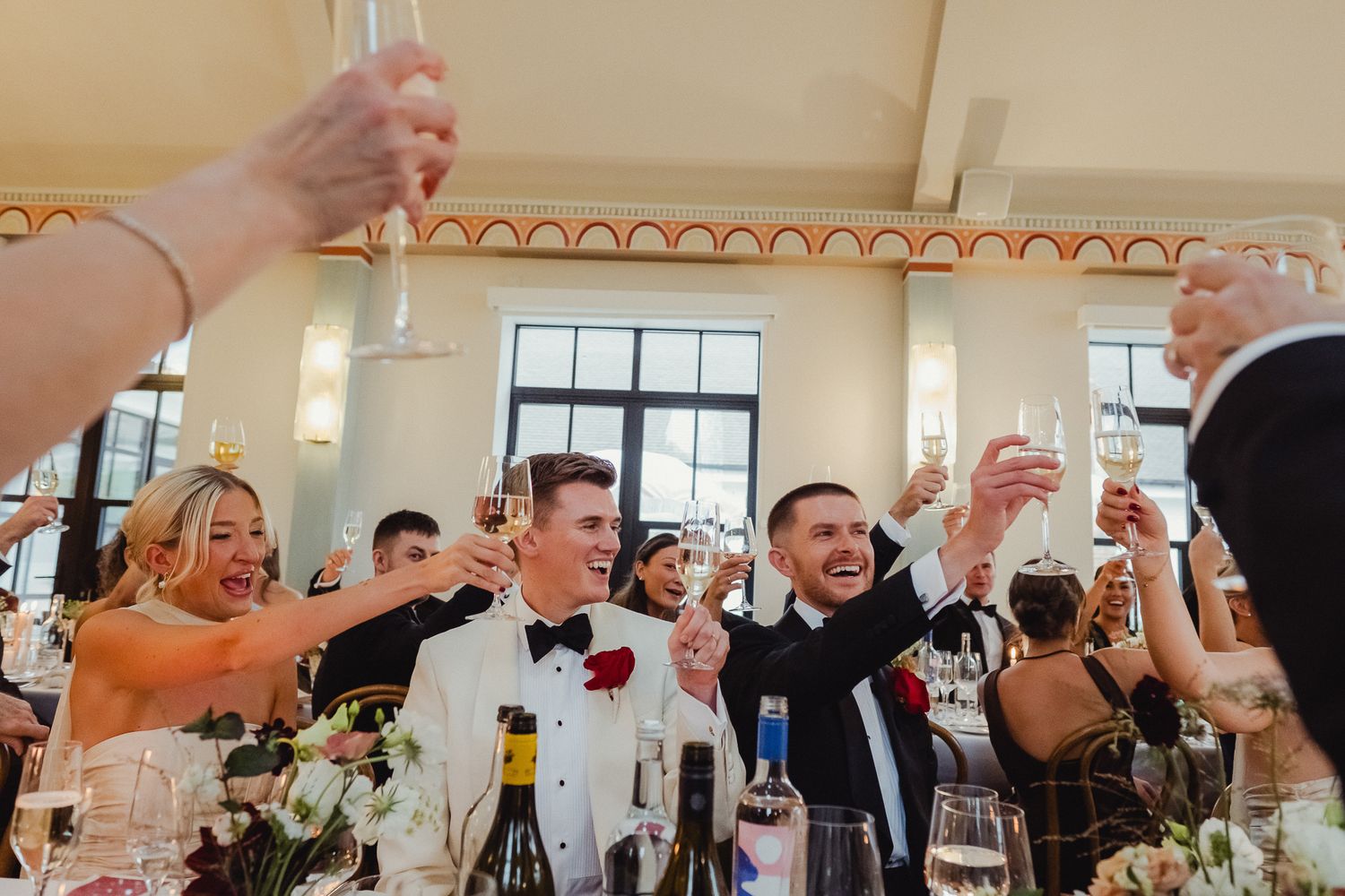 Wedding guests raise their glasses in celebration during a reception toast at an indoor venue.