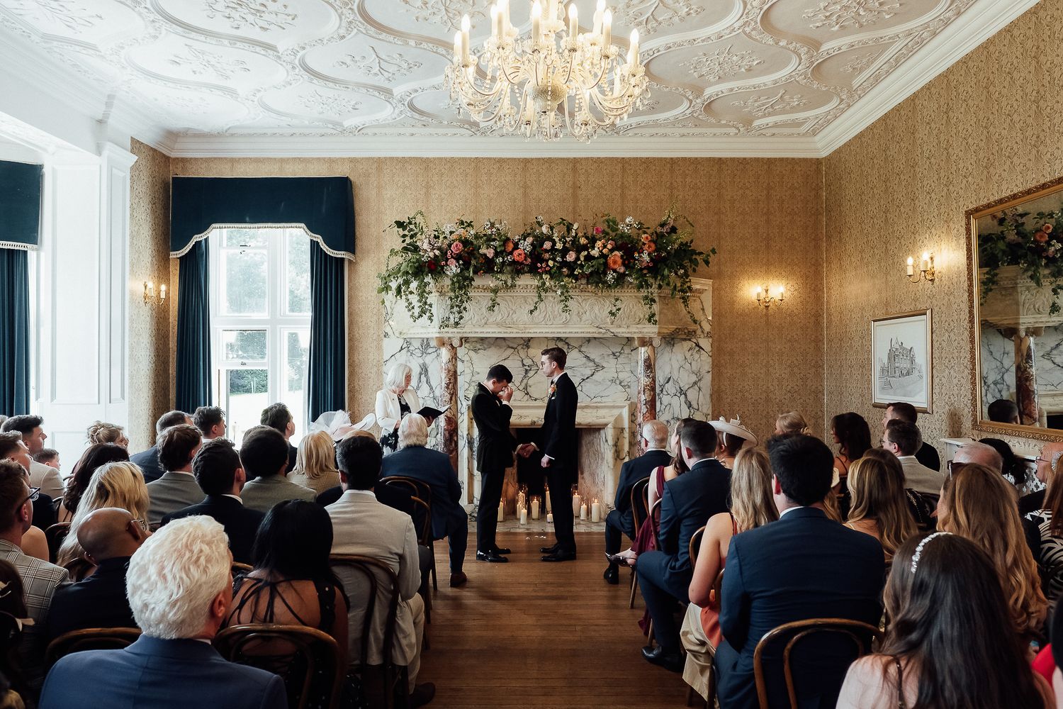 Elegant wedding ceremony in ornate room with floral fireplace arch, chandelier, and seated guests witnessing couple exchange vows.