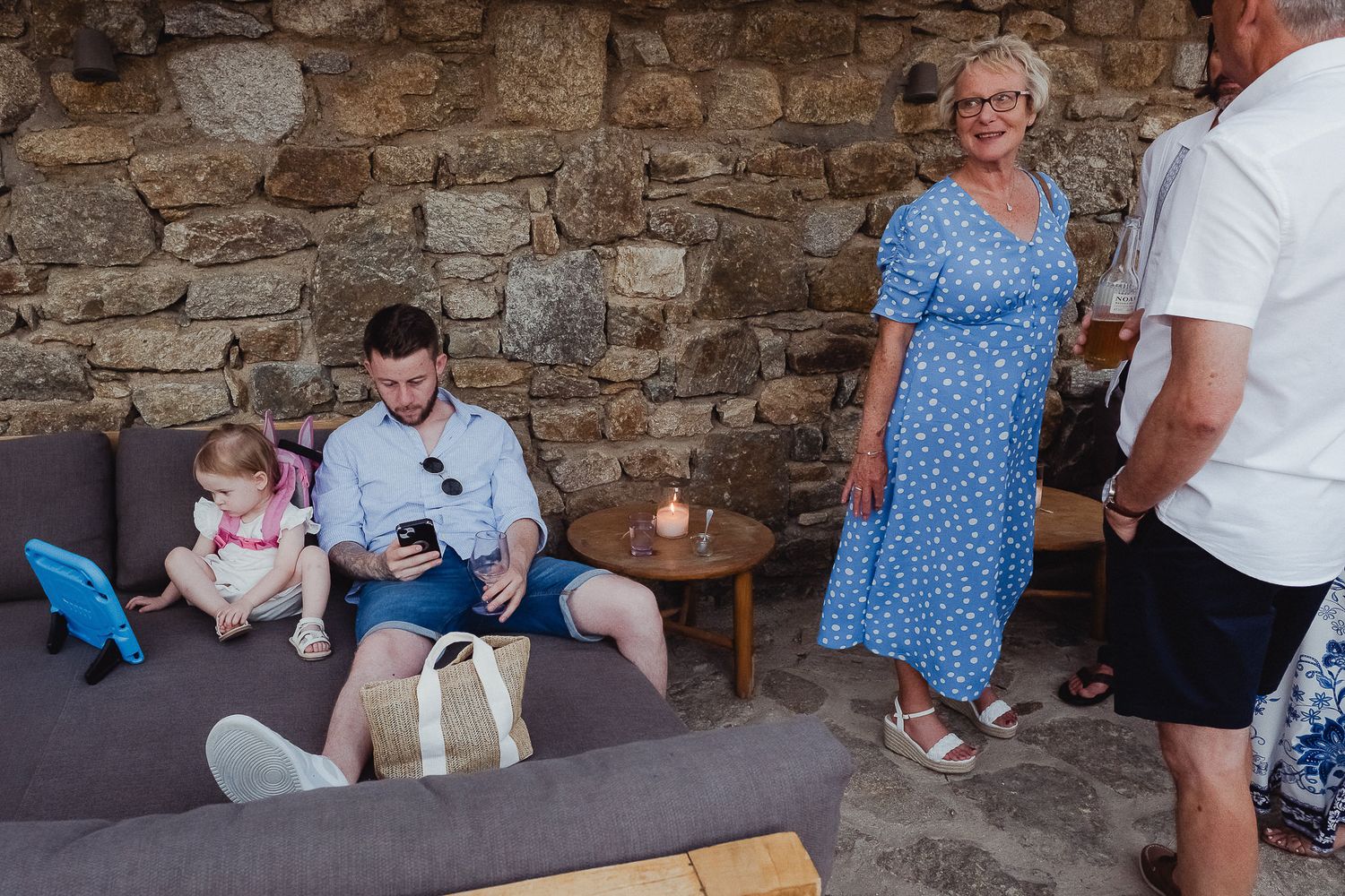 Family gathering on outdoor patio with stone wall backdrop as people relax on couch and stand nearby.