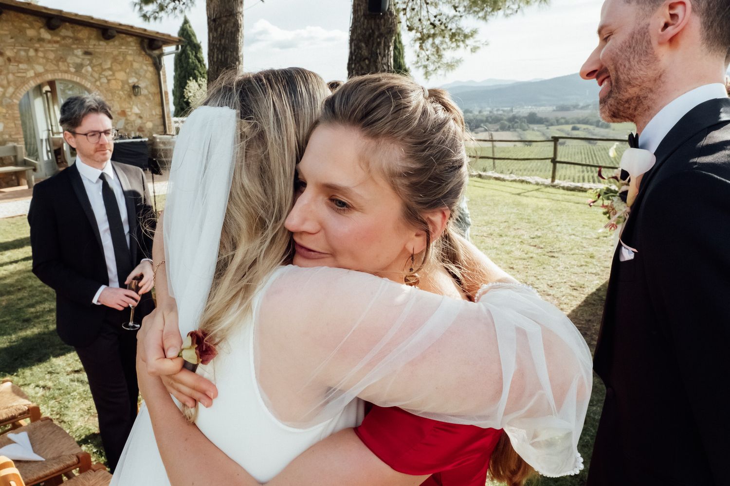 Bride in a white veil embraces a woman in a red dress at an outdoor Tuscan wedding while a groom and guest look on.