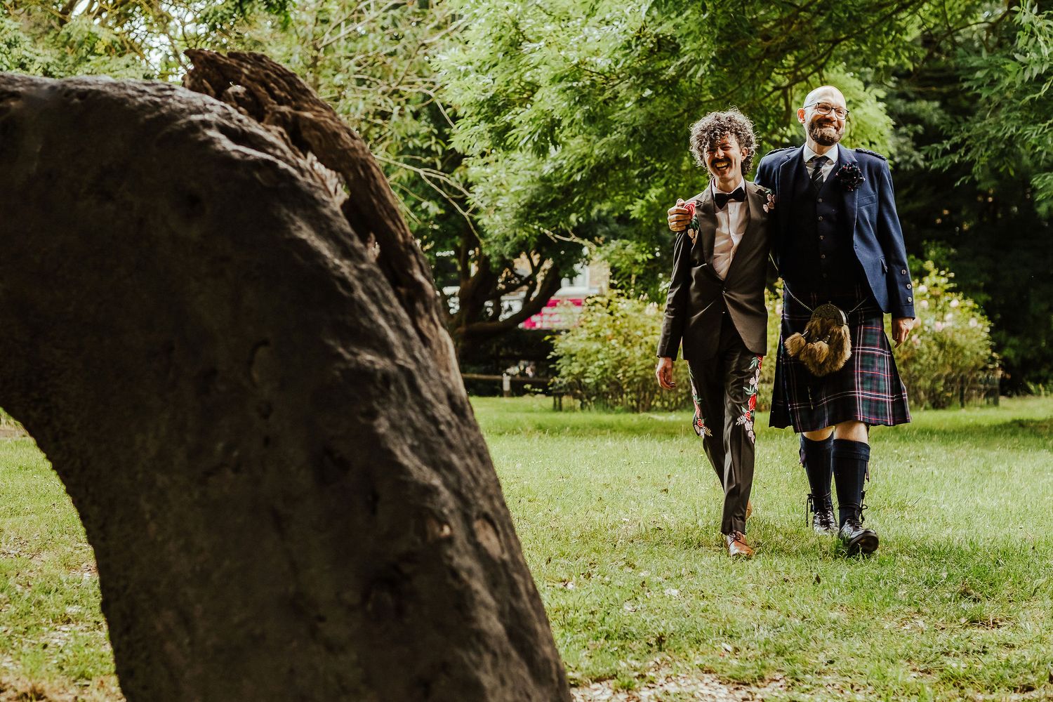 Two smiling people in formal Scottish attire, one in a kilt, walk hand in hand across a sunny green lawn past a large tree trunk.