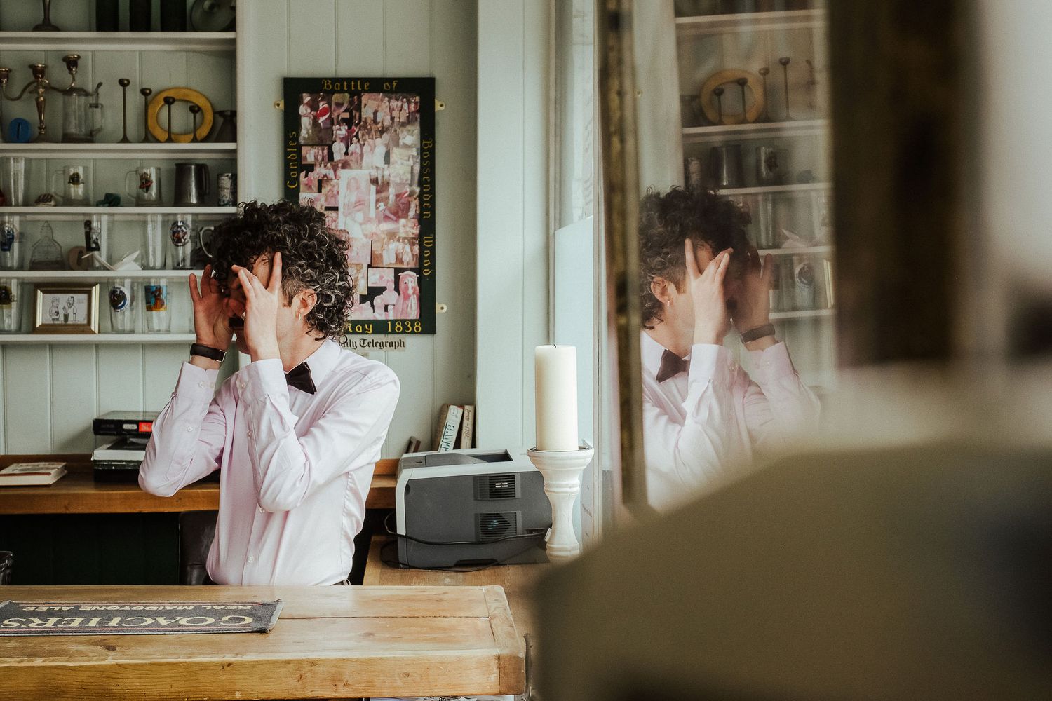 Man in a white shirt and bow tie covers his face with his hands in a rustic room with shelves, framed photos and a reflection in a mirror.