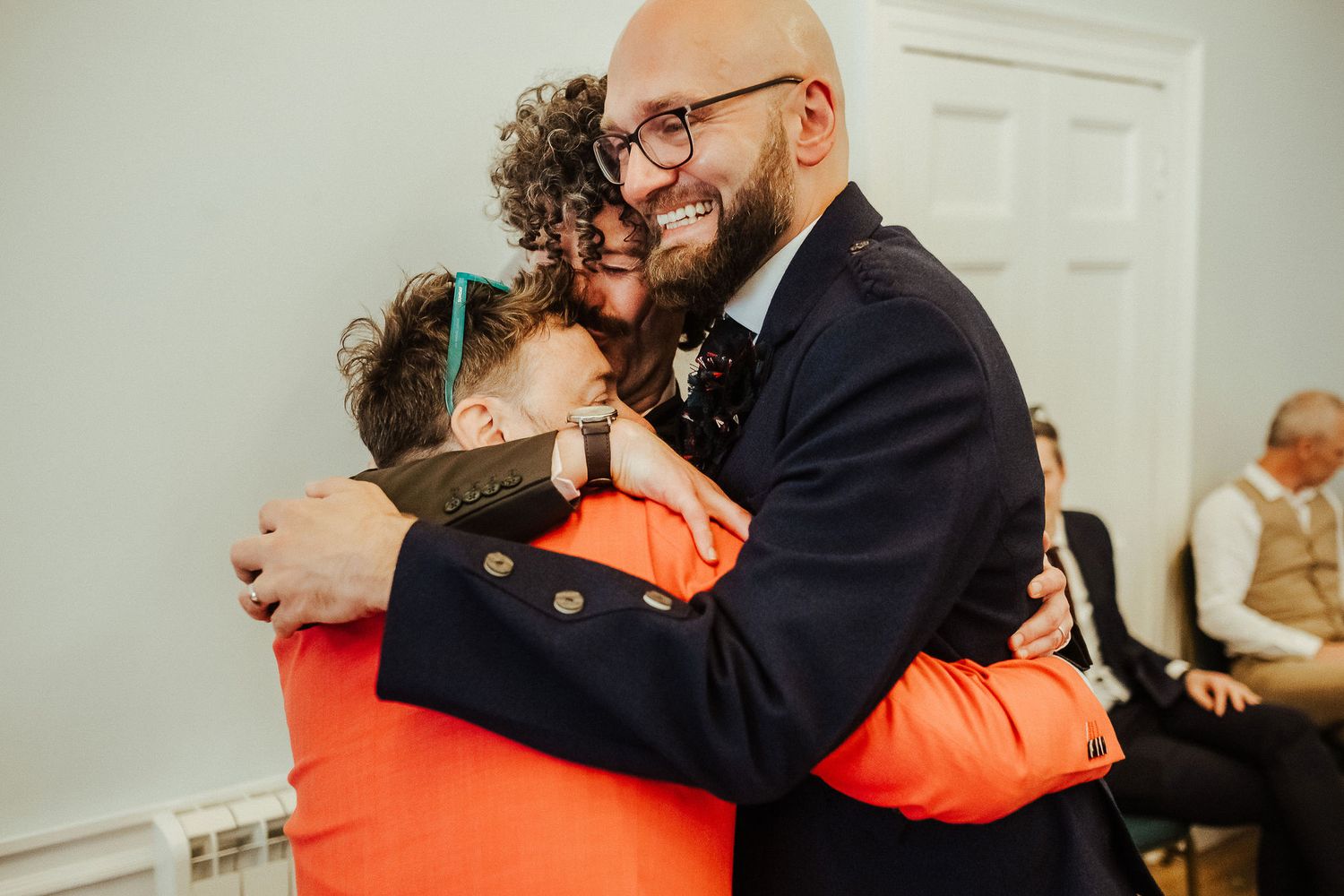 A bearded man in glasses and a navy blazer hugs a person in an orange top warmly while guests sit in the background.