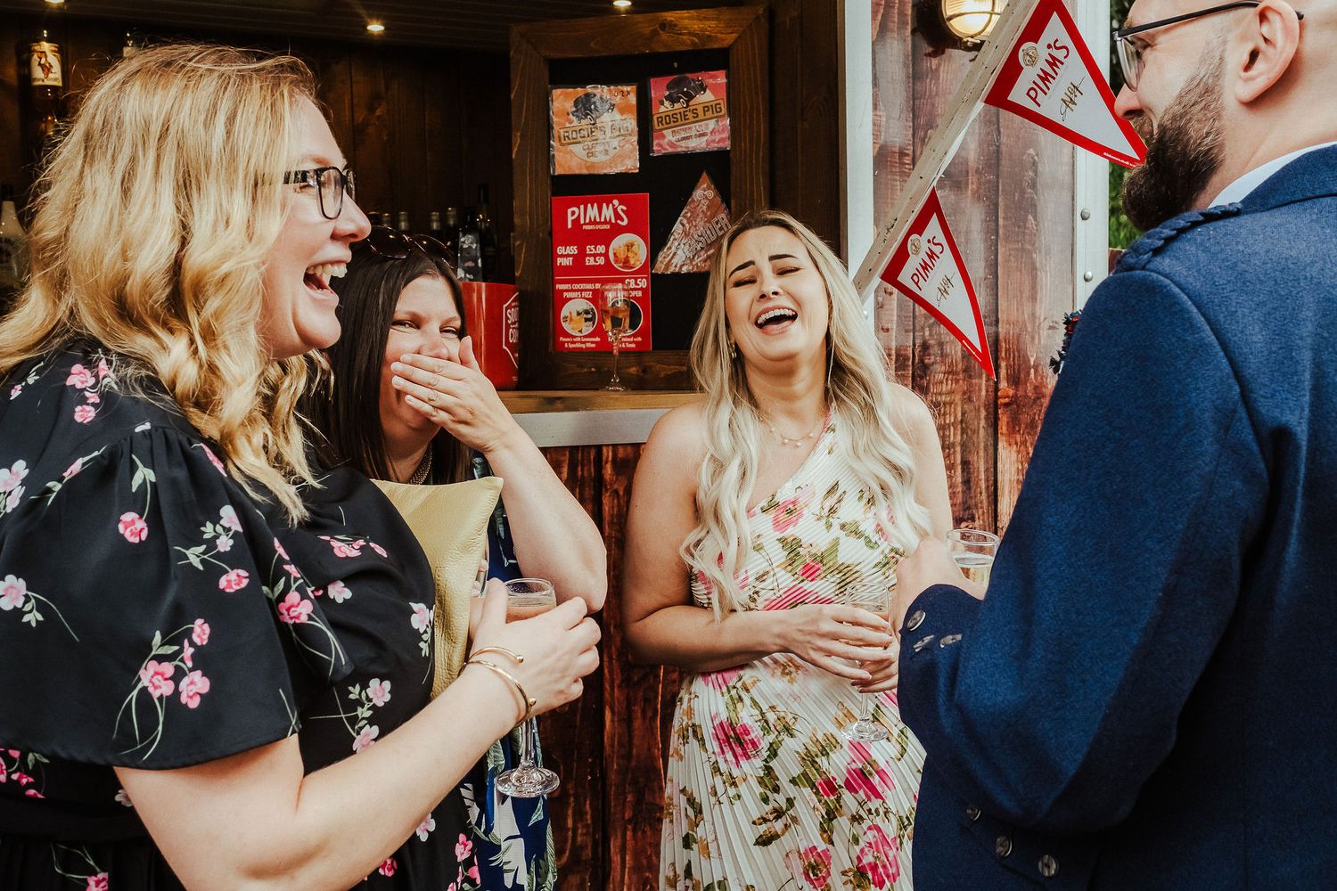 Group of laughing friends in floral dresses chatting and enjoying drinks at a lively indoor social gathering with a man in a blue jacket.