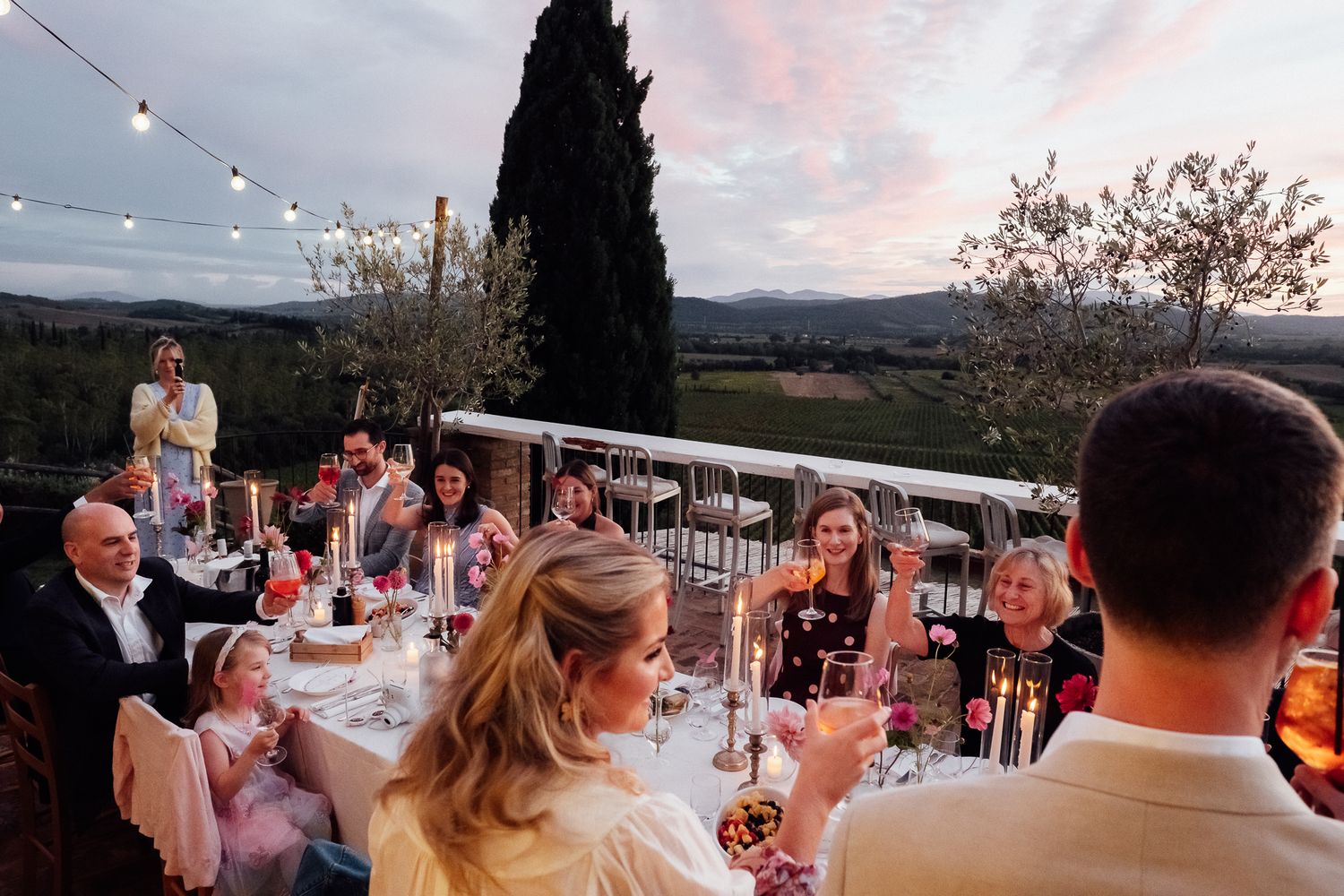 Family and friends gather around a long candlelit dinner table at an outdoor celebration with string lights and rolling hills at sunset.