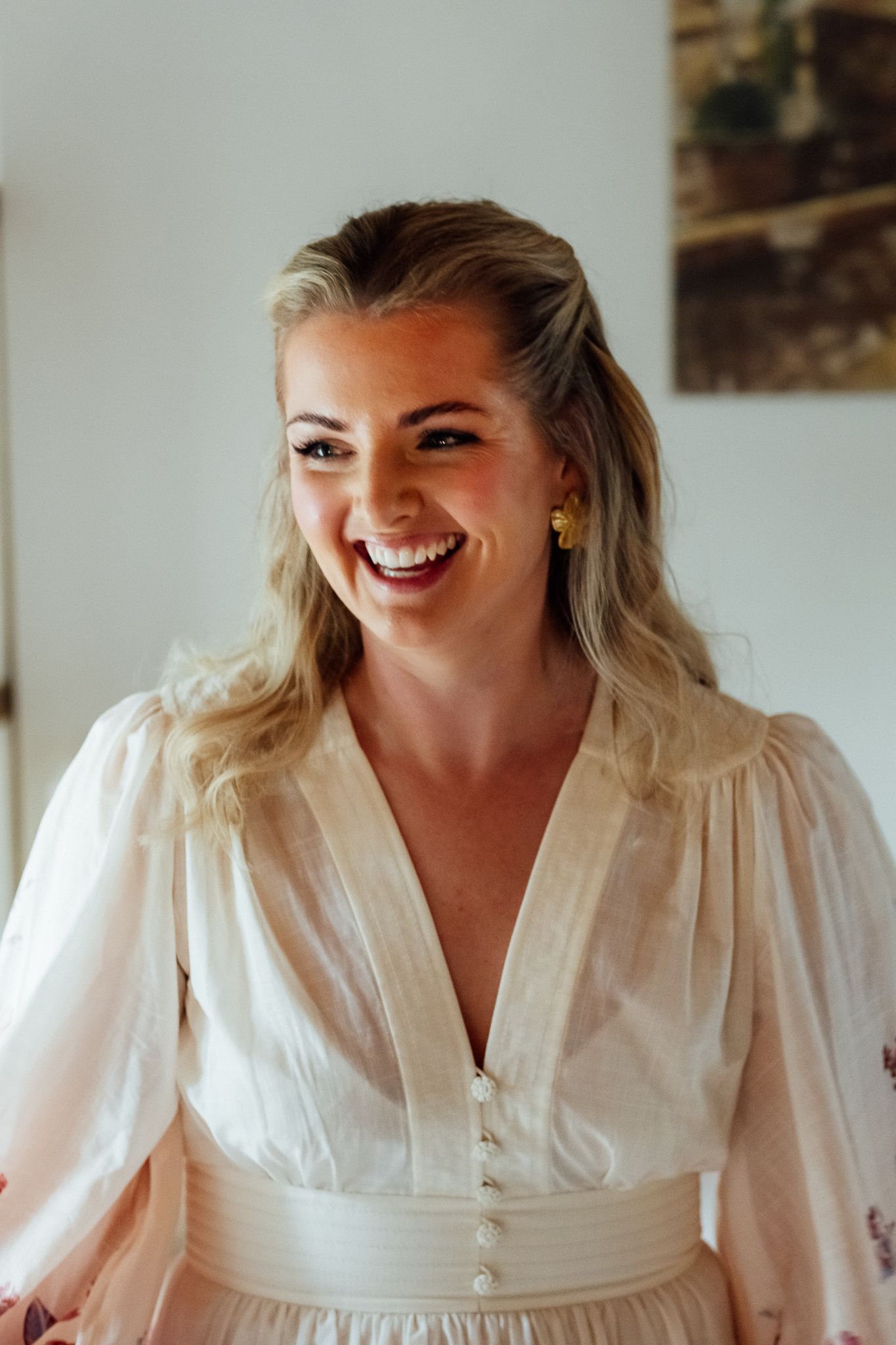 Smiling woman with wavy blonde hair in a half-up style wears a cream puff-sleeve dress with a button front and gold earrings.