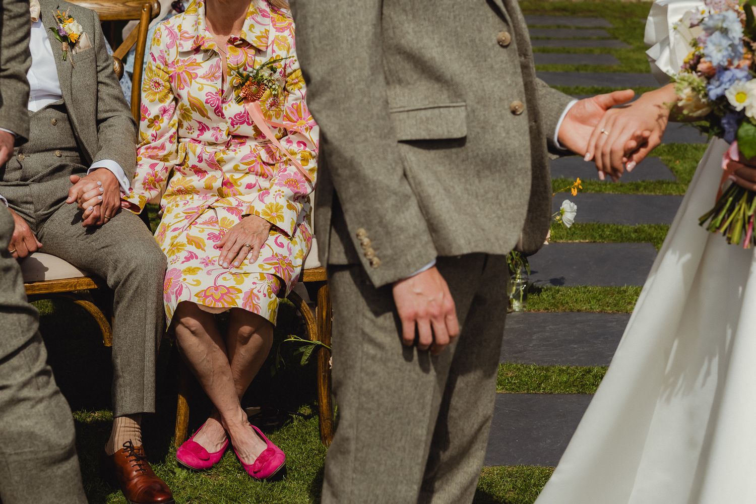 Wedding party members in gray suits and pink floral bouquets pose together outdoors on grass.