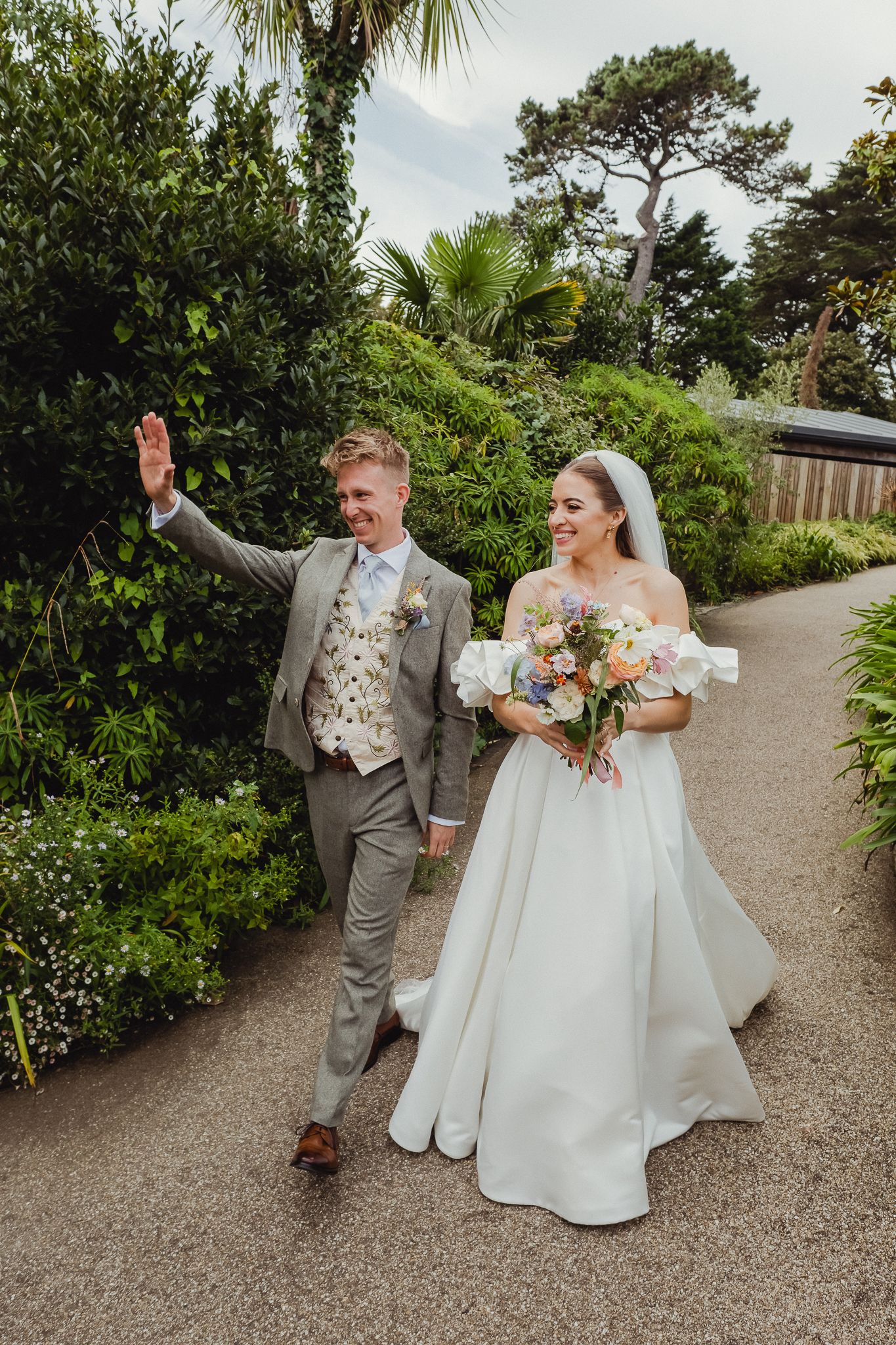 A couple in wedding attire dances and celebrates joyfully on a garden path surrounded by lush greenery and palm trees.