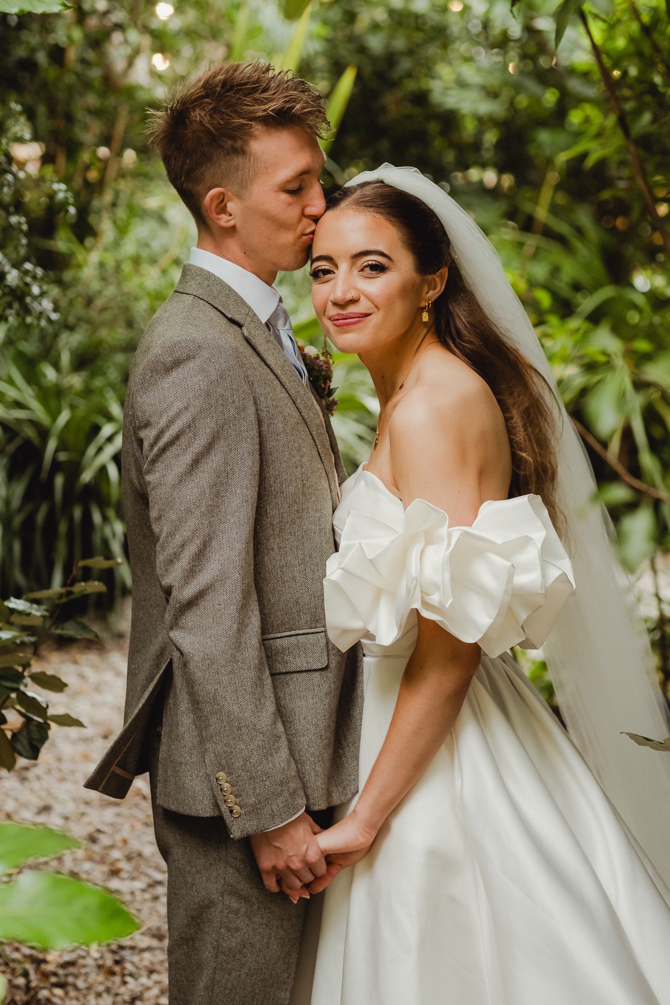 Romantic wedding couple poses together in a lush garden setting wearing a white gown and gray suit.