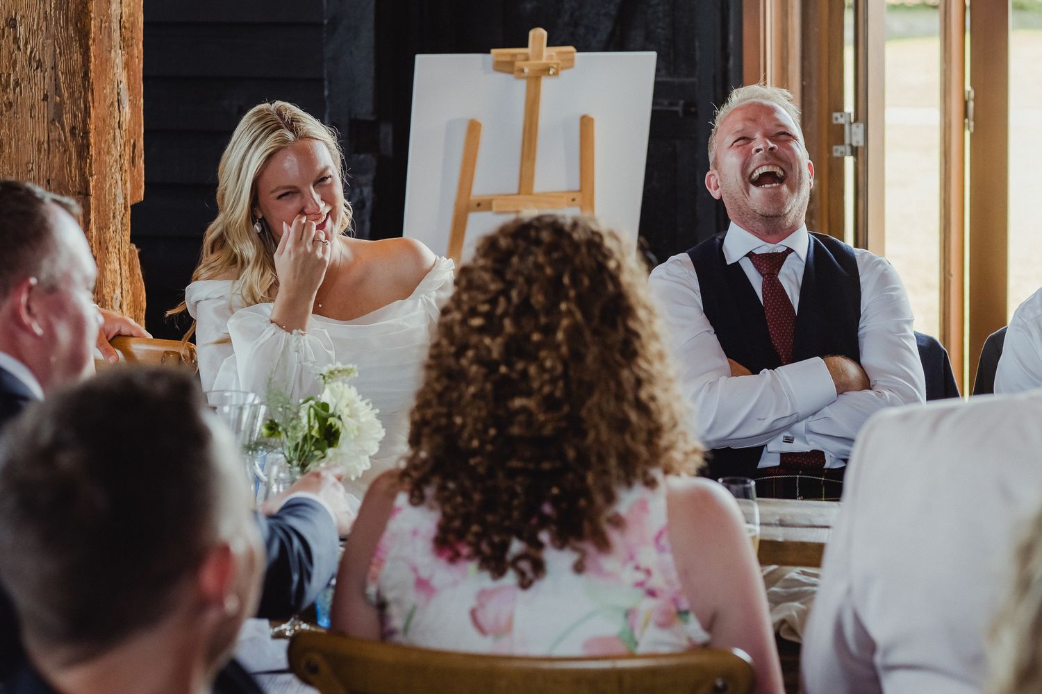 Wedding guests laugh and smile during a reception in a rustic wooden venue with natural lighting.