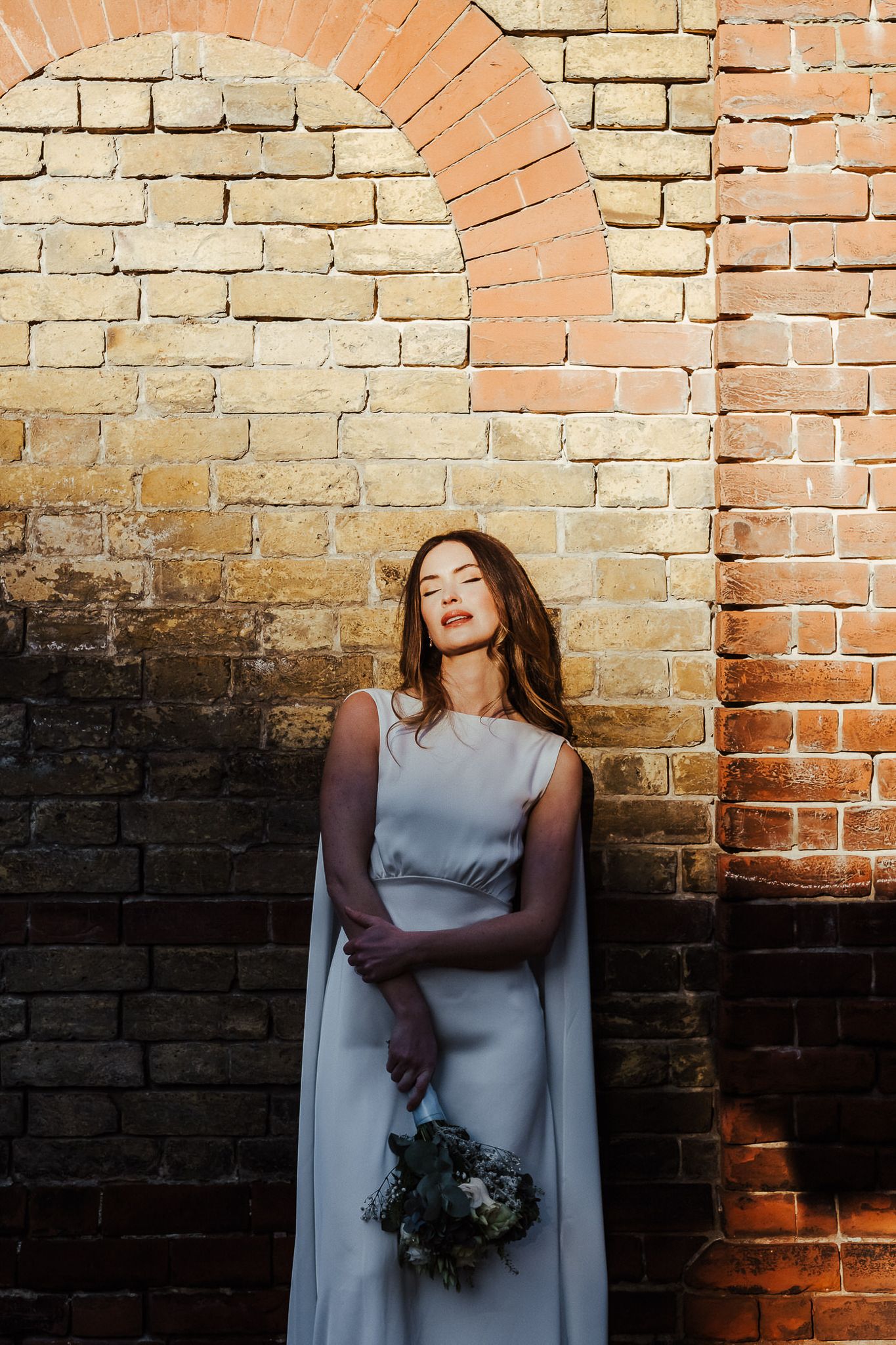 A person in a light blue dress stands against a rustic brick wall holding a dark bouquet of flowers.