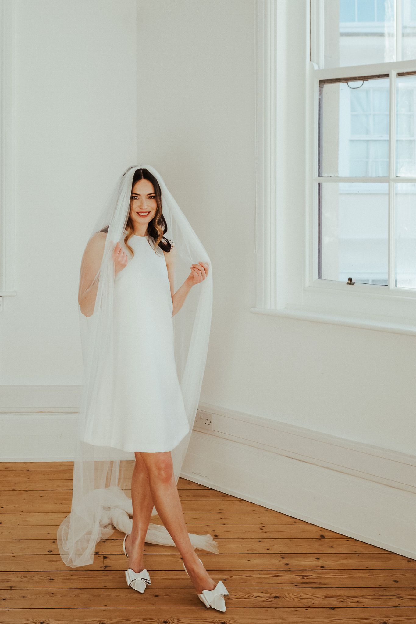 A bride in a short white dress and veil poses playfully in a bright studio with wooden floors and white walls.