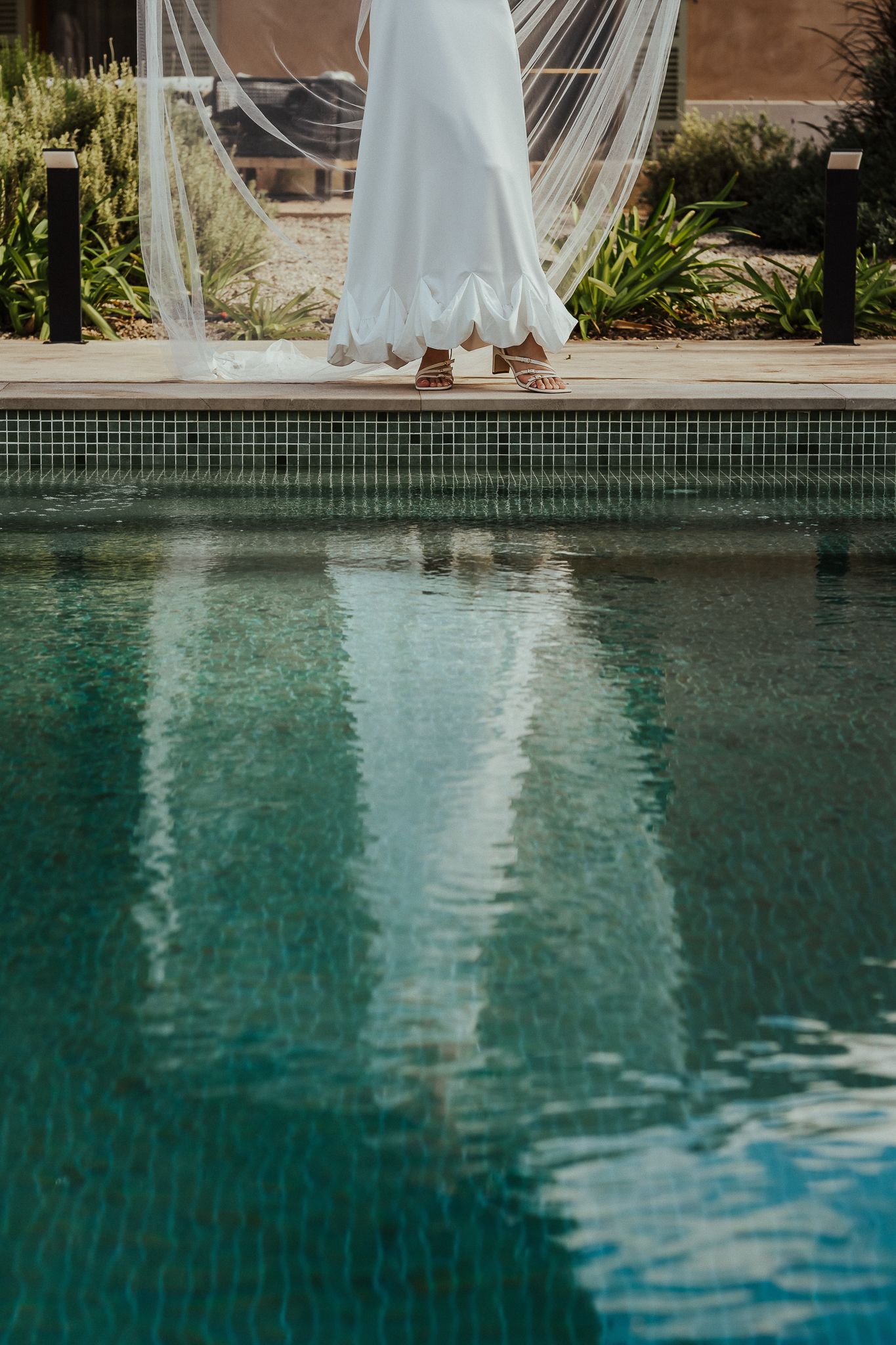 A flowing white wedding dress veil reflects in a serene turquoise swimming pool on a sunny day.