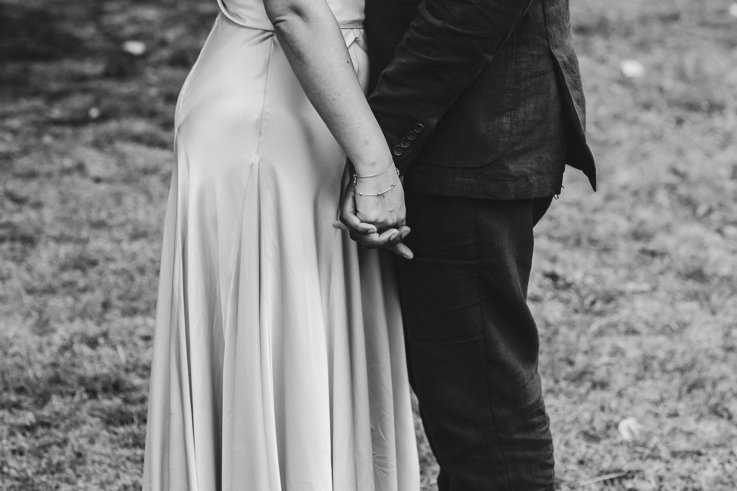 A couple in formal wedding attire holds hands while standing in a rustic outdoor setting in black and white.