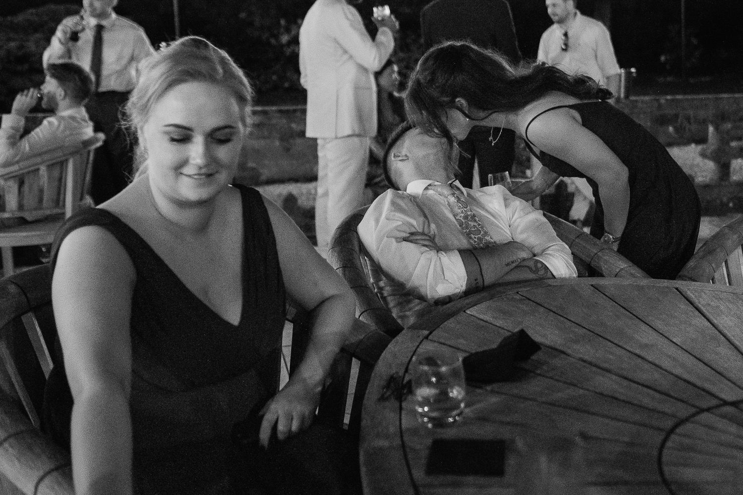 Black and white photo of a woman in a black dress sitting at an outdoor wedding reception event.