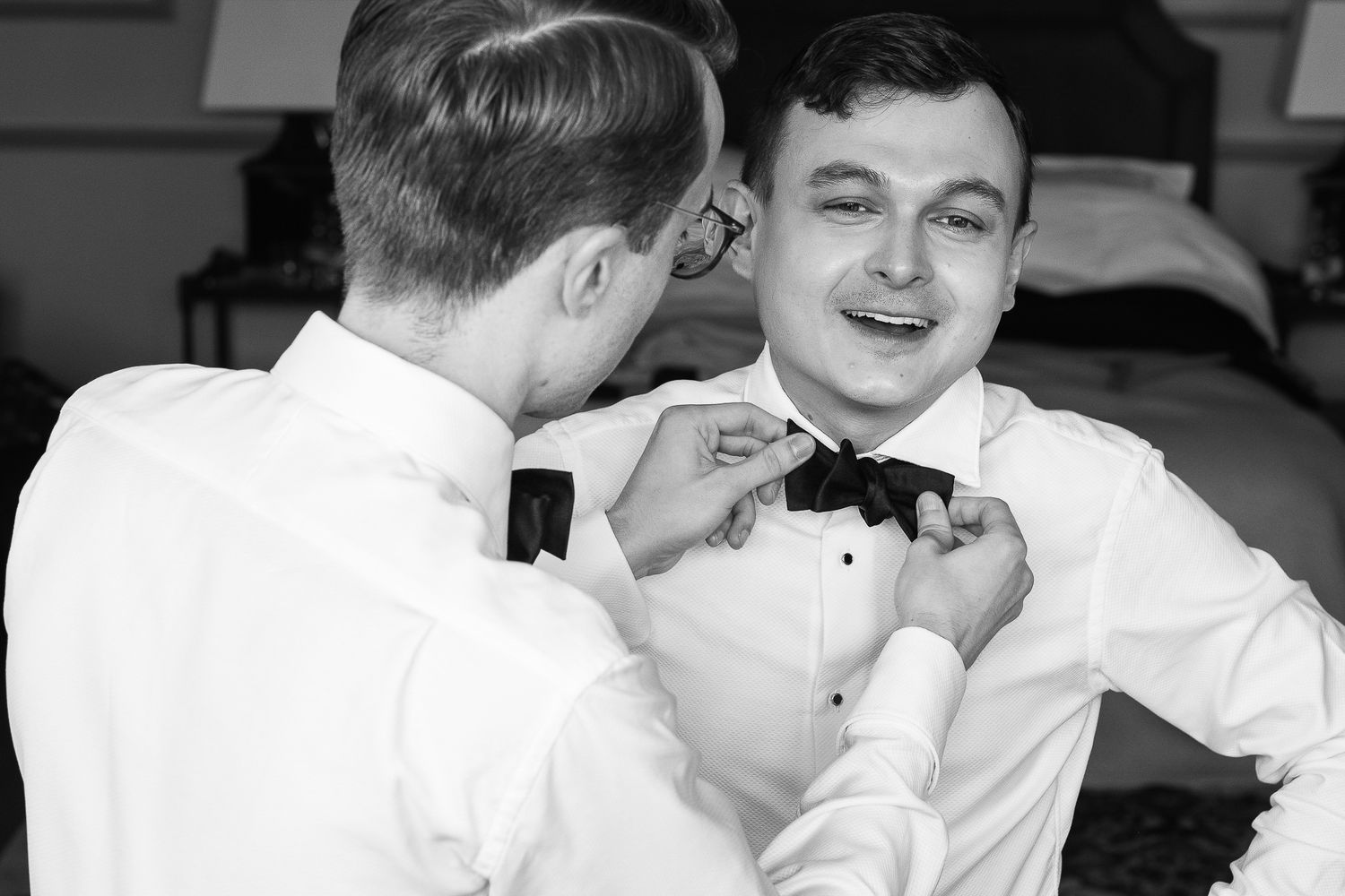 Black and white photo of two men in white dress shirts adjusting a black bow tie before a wedding ceremony.
