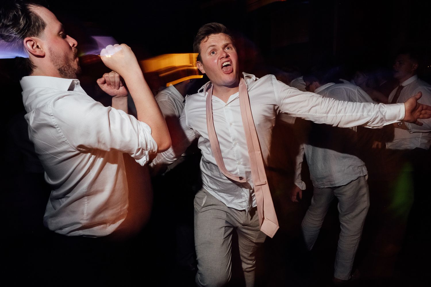 Men in white shirts dancing energetically at a wedding reception party under dark atmospheric lighting.