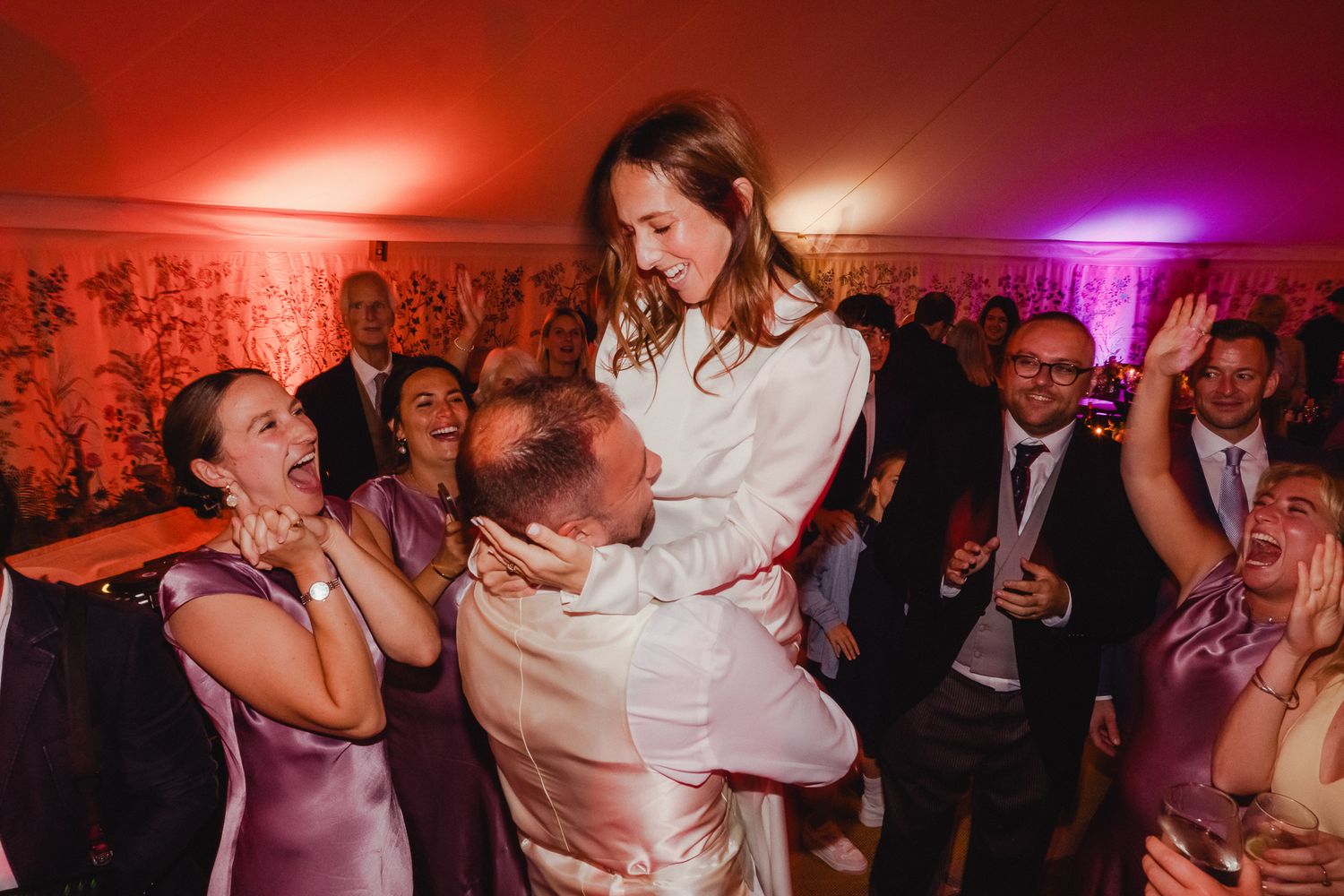 Wedding guests dancing and celebrating on a dimly lit dance floor with red and purple lighting effects.