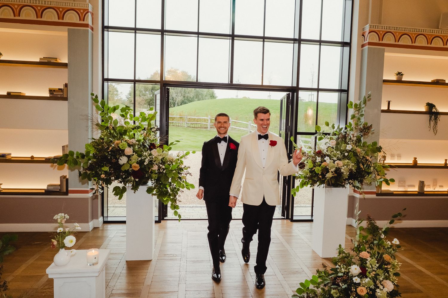 Two people in formal attire walk down a bright hallway decorated with potted plants and white shelving.