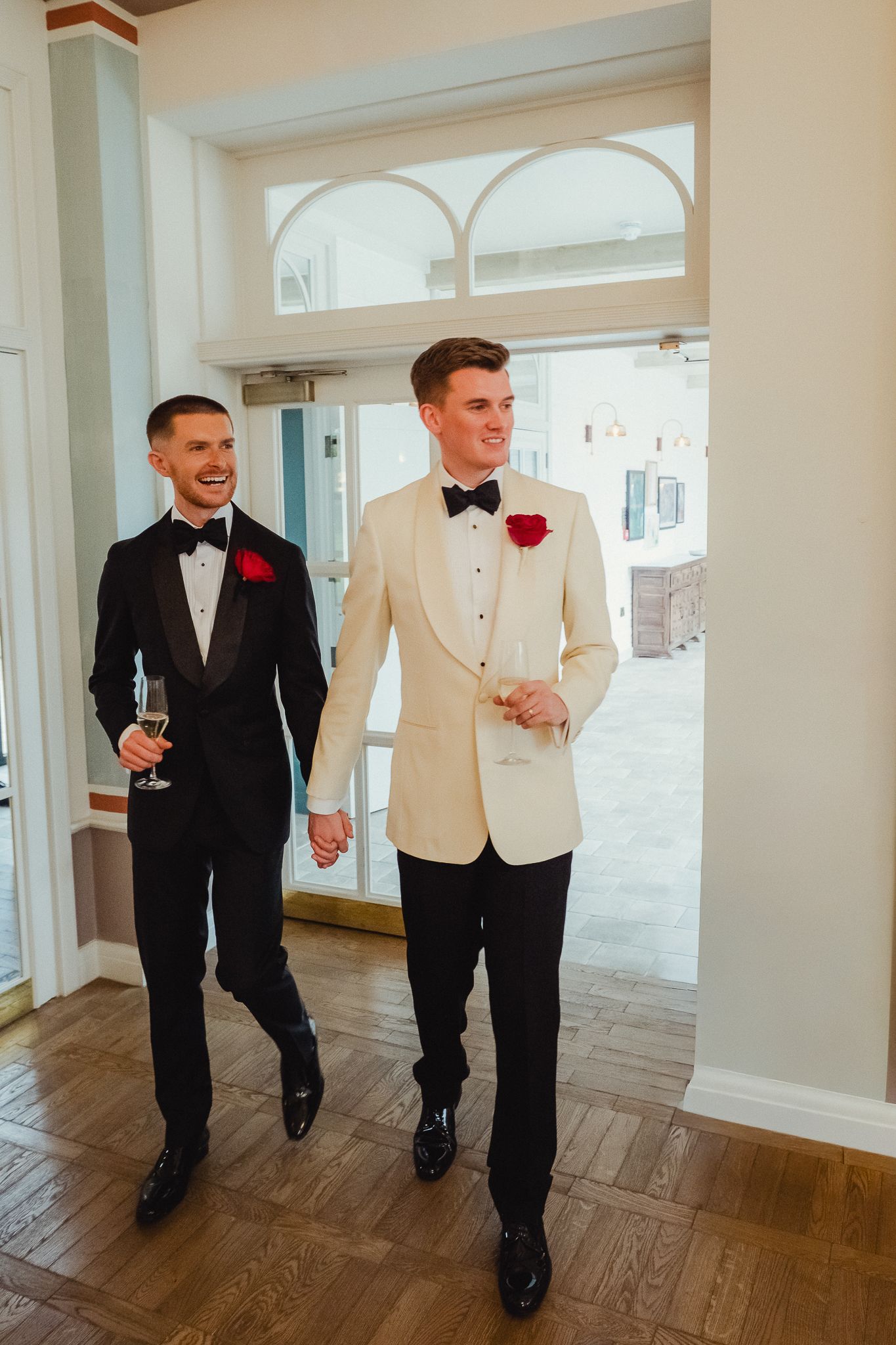 Groomsmen in black tuxedos and white dinner jackets walk and laugh together on hardwood floors at a wedding venue.