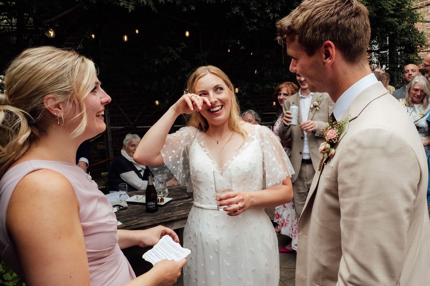 A smiling bride in a white lace dress wipes a happy tear as she stands with a groom and a woman at an outdoor wedding celebration.