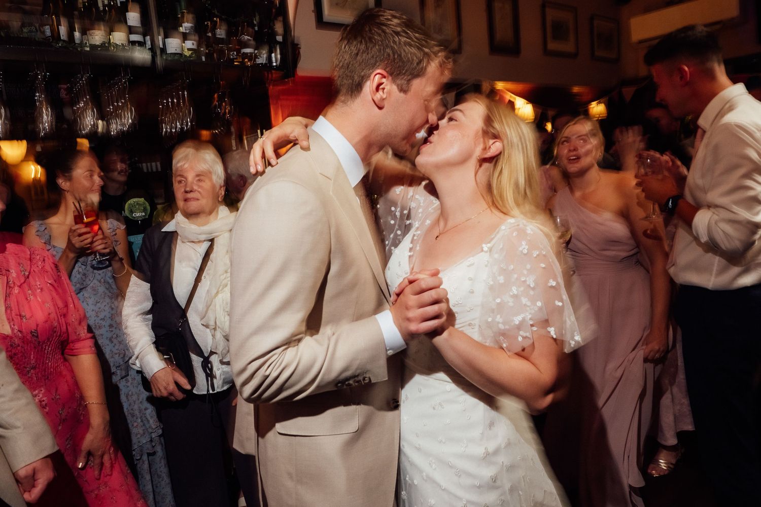 A bride in a white lace dress and a groom in a beige suit share a close dance at a lively wedding reception while guests look on.