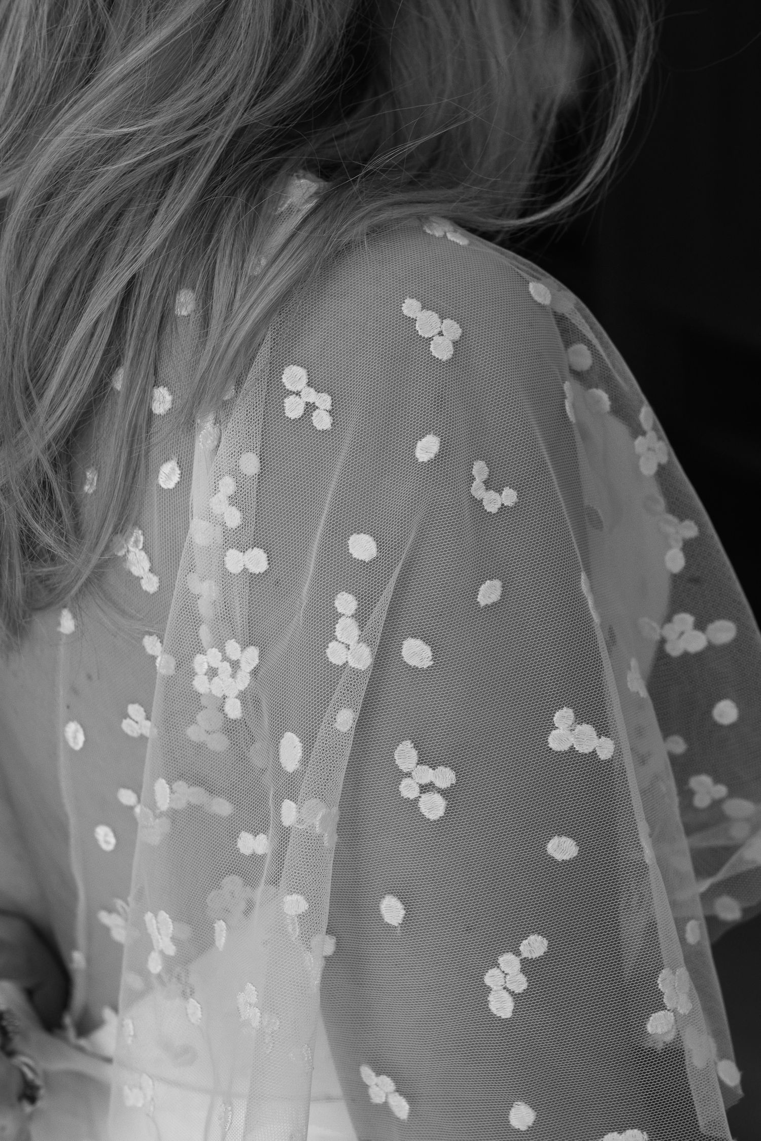 Black and white photo of a woman from behind with long hair, wearing a sheer tulle garment with scattered floral appliqué details.
