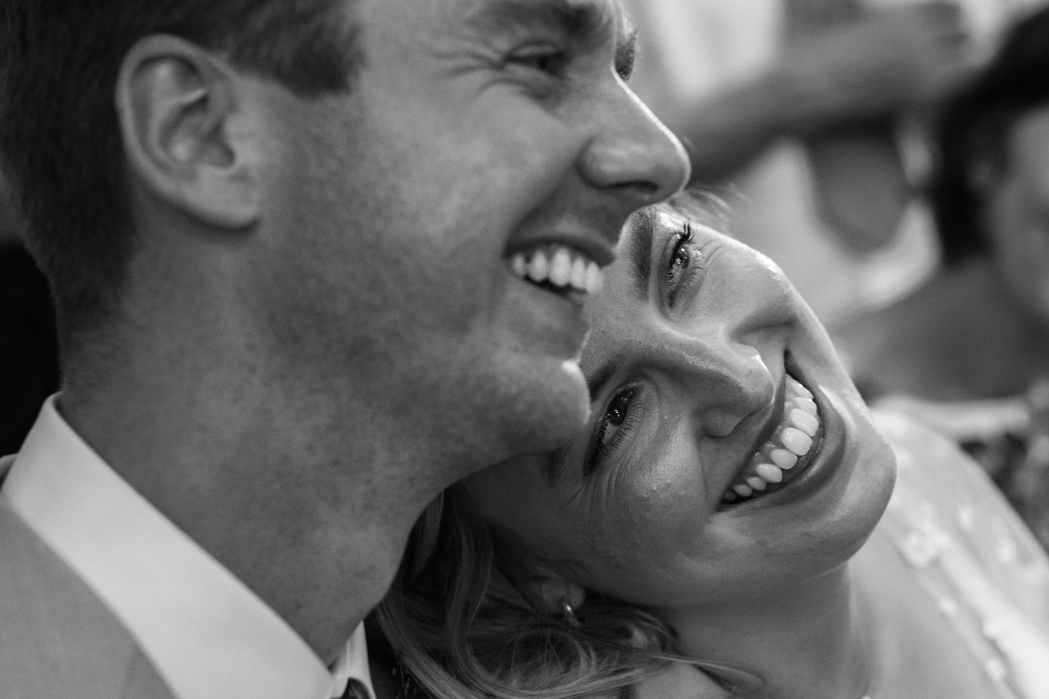 Black and white close-up of a smiling man and woman laughing joyfully together, with her head resting against his shoulder.