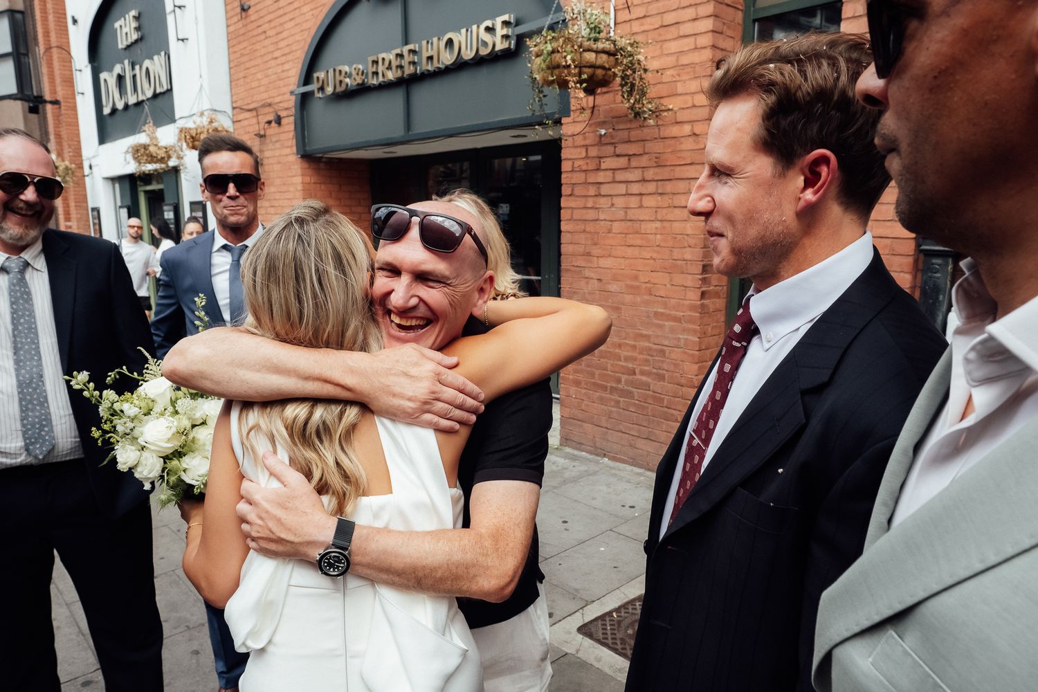 A bride in a white dress warmly hugs a smiling man in sunglasses outside a brick building while groomsmen in suits look on.