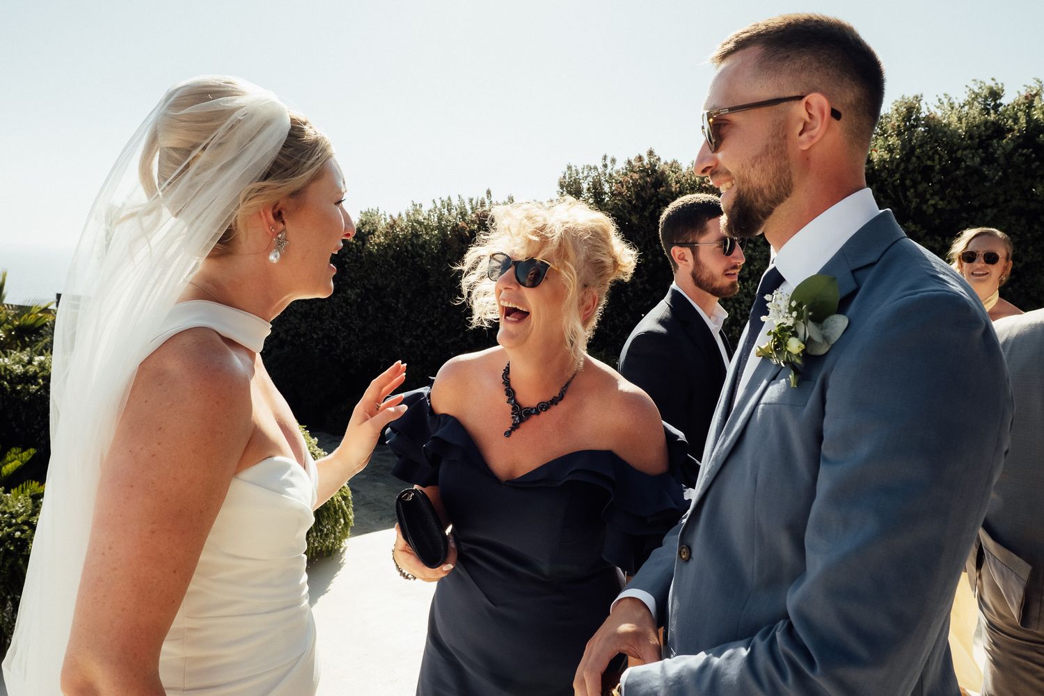 Bride and groom share a joyful moment with a guest at an outdoor wedding reception on a sunny day.