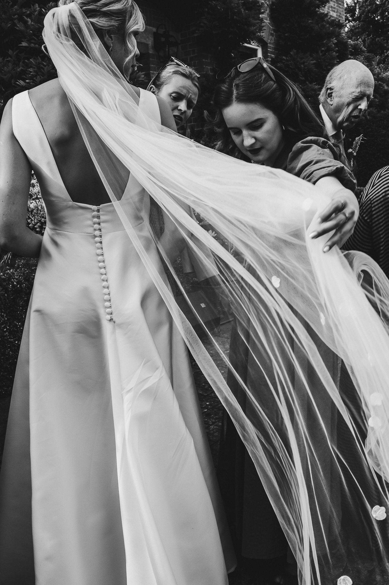 A close-up black and white shot of guests admiring a bride's flowing veil outdoors.