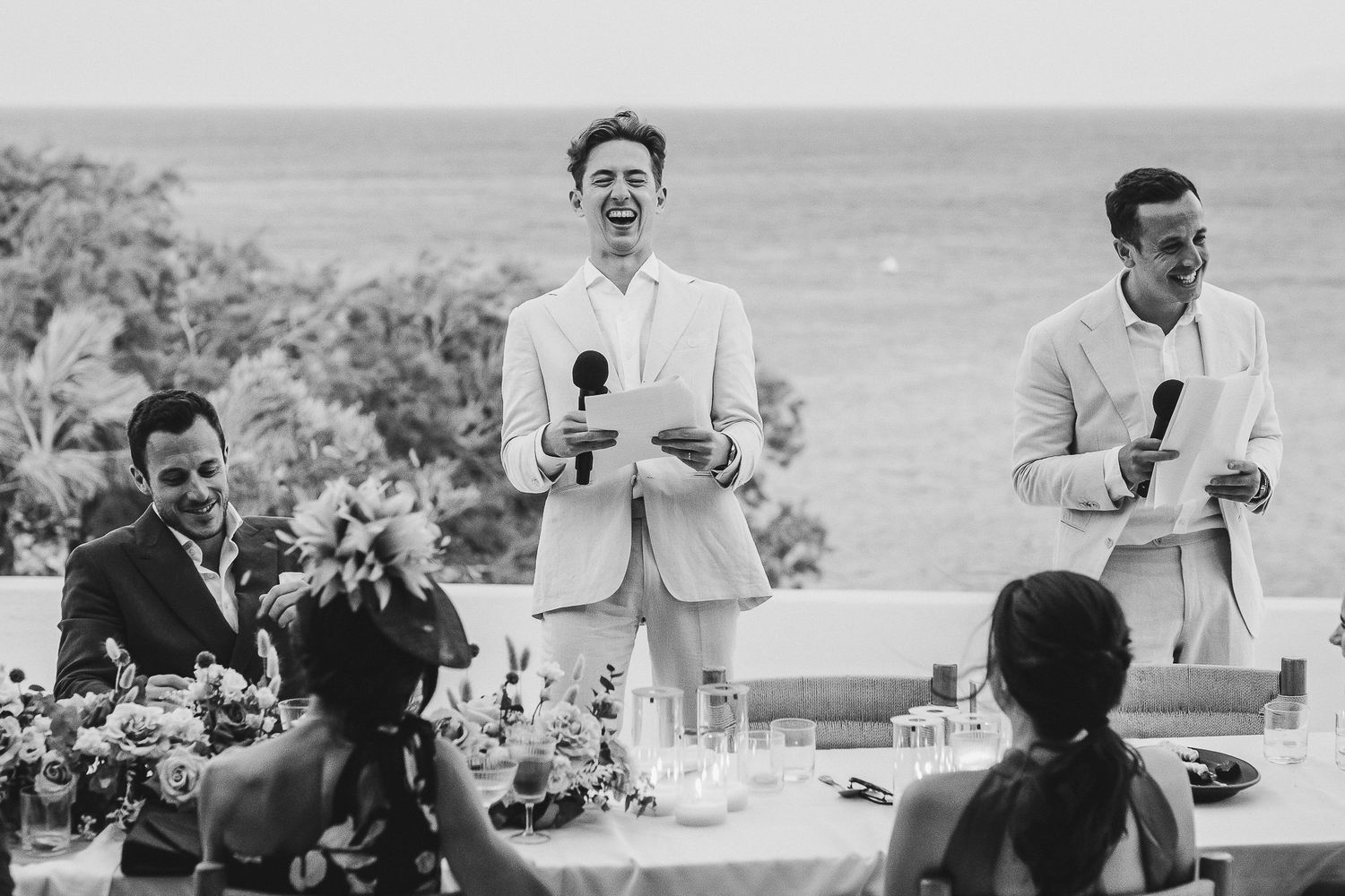 Black and white image showing joyful reactions and laughter during wedding speeches at an outdoor reception.