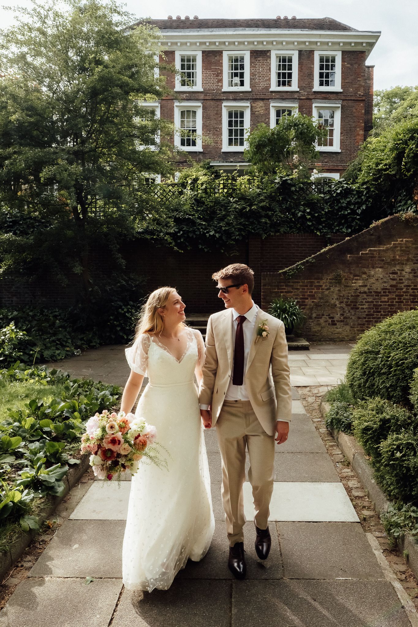 Bride and groom walk hand-in-hand through a lush garden path, with a classic brick manor house in the background.