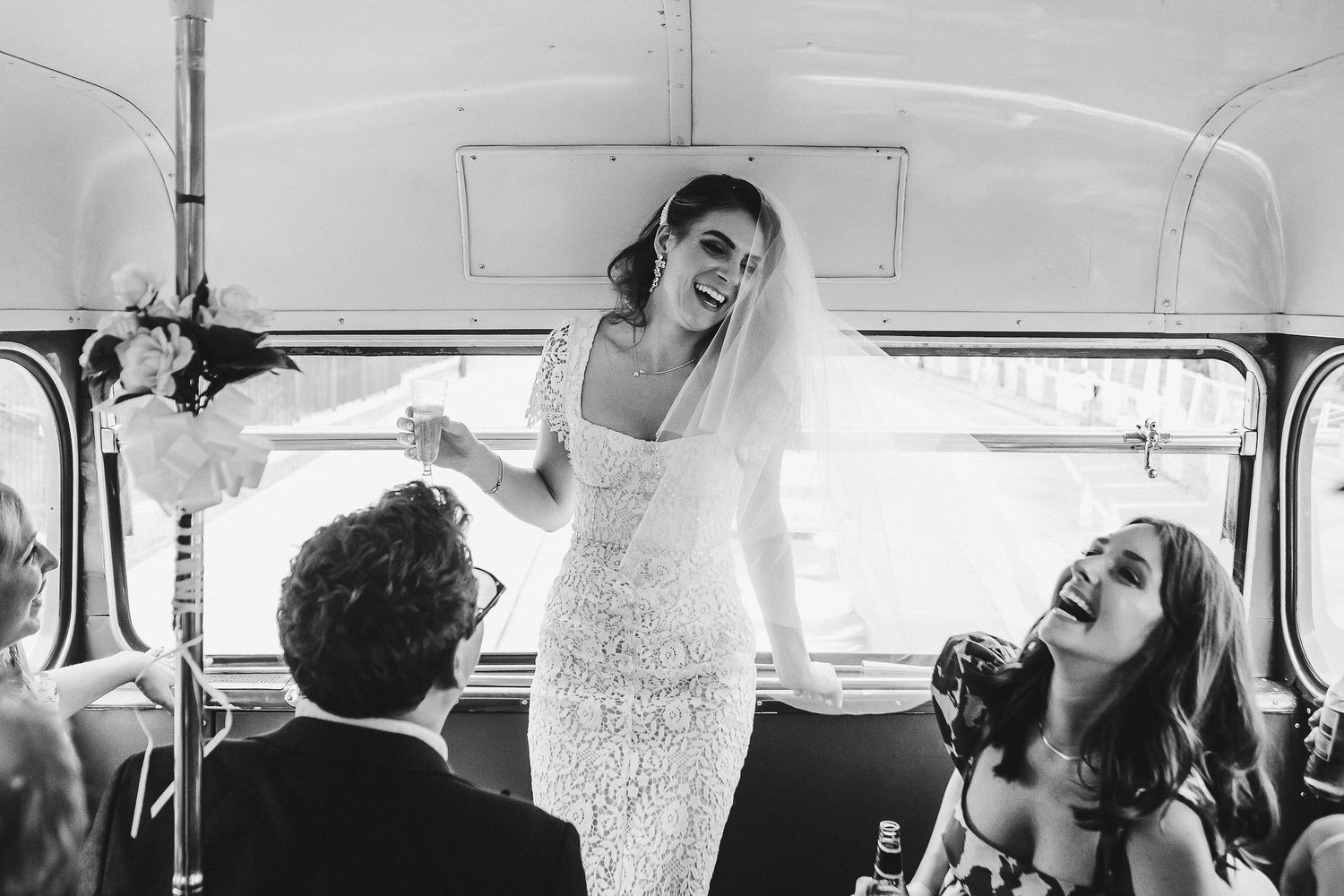 A joyful moment captured in black and white inside a vintage bus with a bride in a lace dress celebrating with companions.