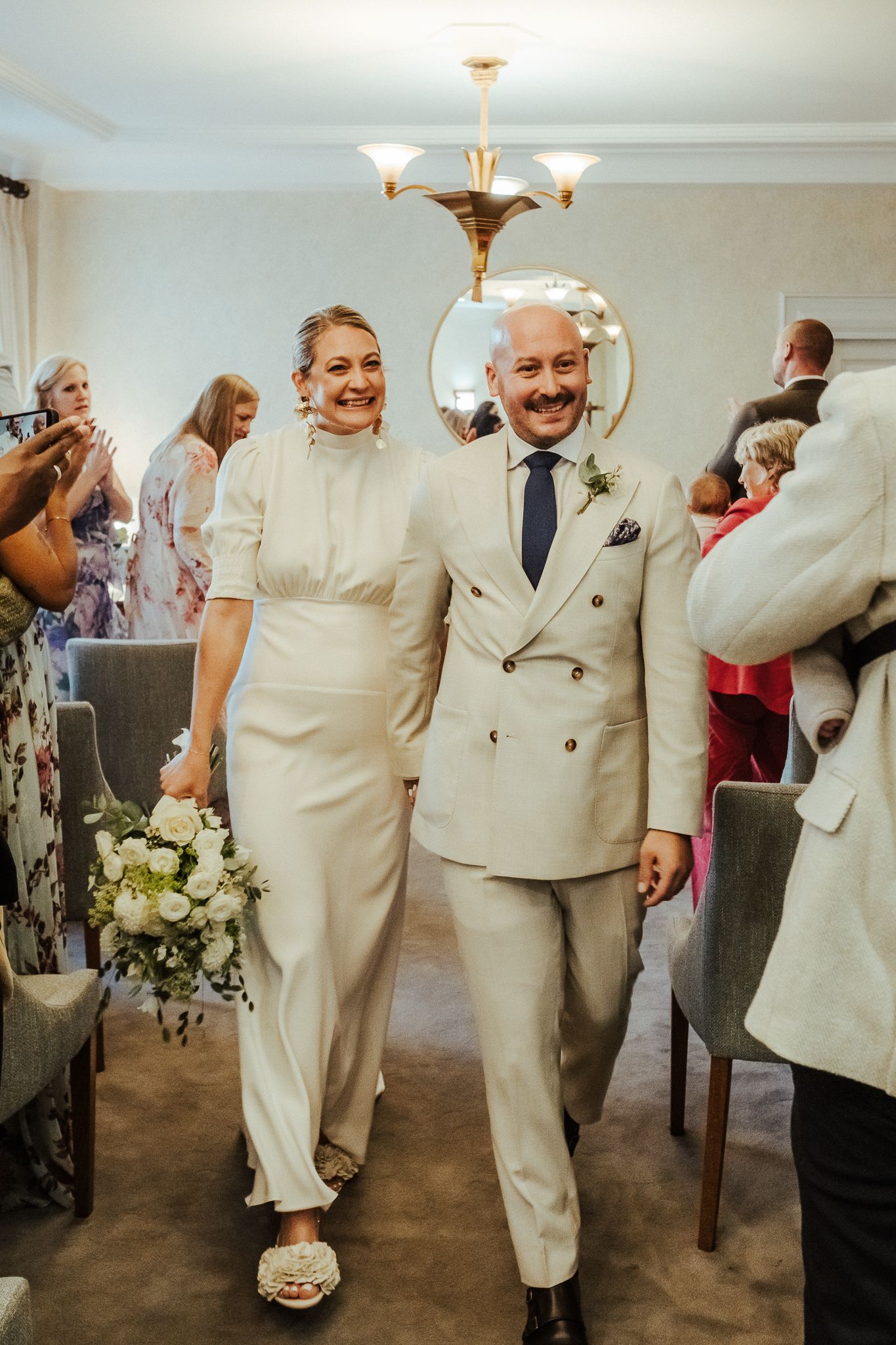 A couple in matching cream-colored formal attire walk together at an indoor celebration while guests applaud.