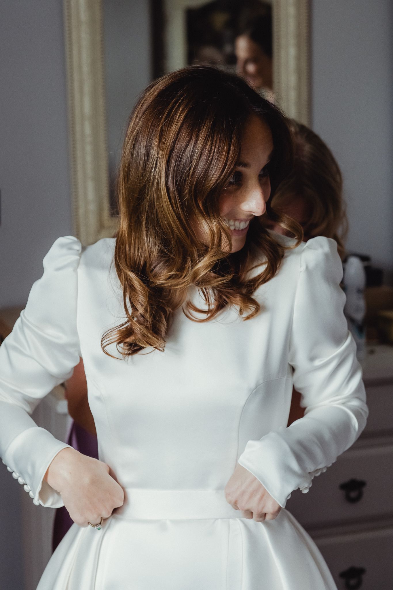 A bride in an elegant white long-sleeved wedding dress with puff sleeves adjusts her gown while looking down.