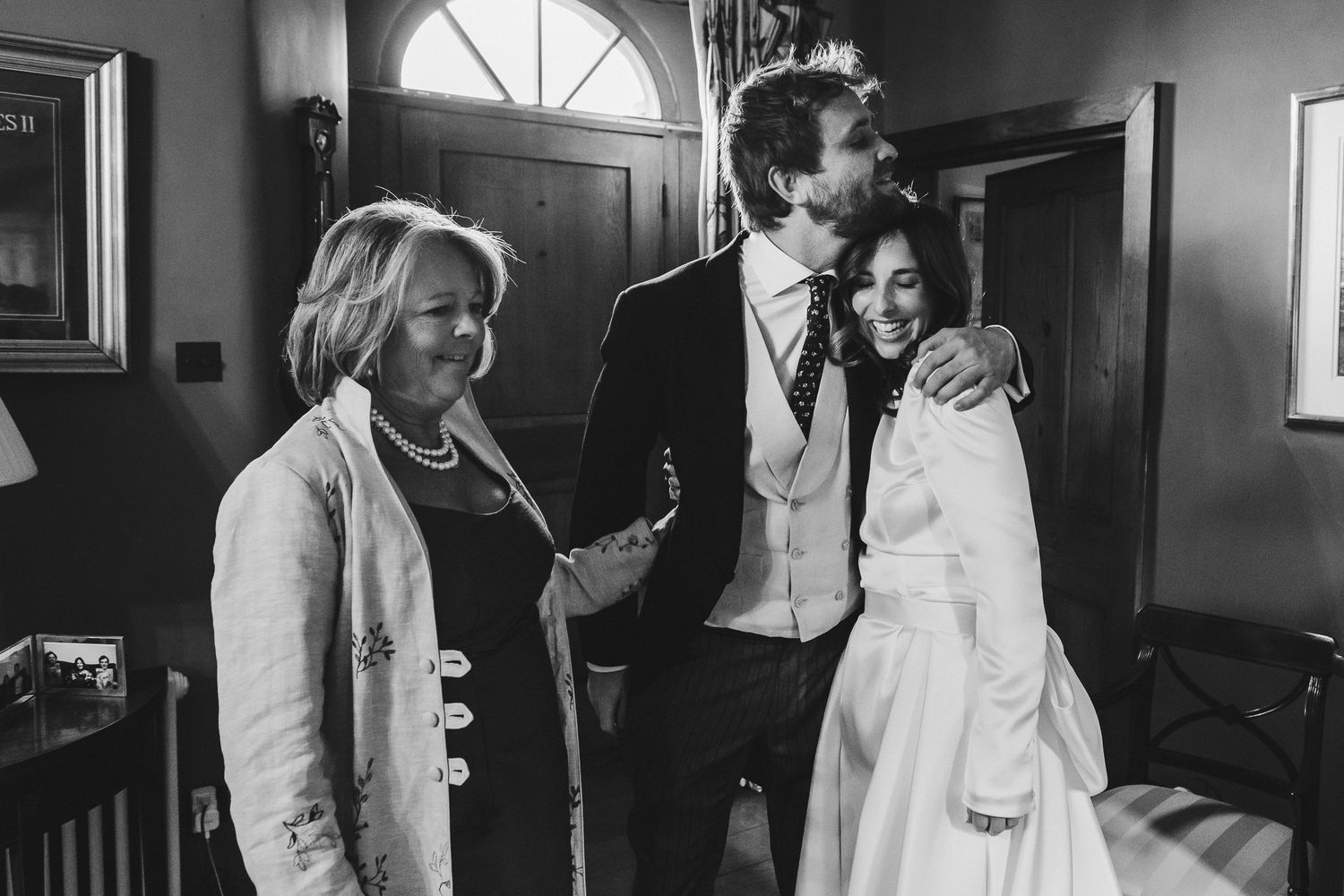 A black and white candid moment captured at an indoor wedding venue showing three people in formal attire.
