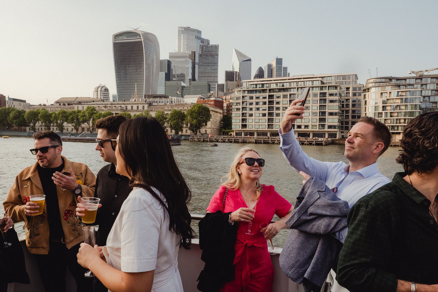People enjoying drinks and views on a boat tour along a city waterfront with skyscrapers in the background.