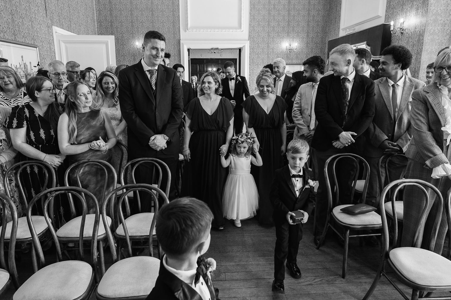 Black and white wedding processional photo with flower girl, ring bearer, and guests in elegant hall.