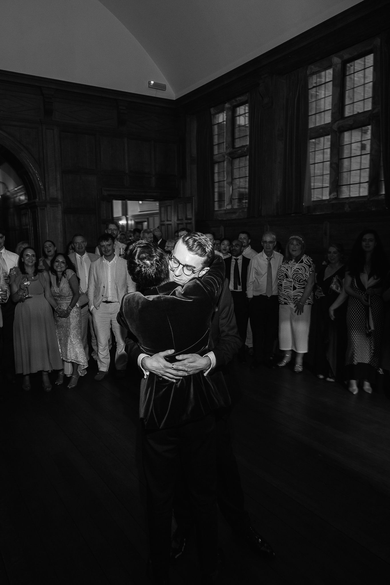 Black and white photo of couple sharing first dance embrace at wedding reception with guests watching.