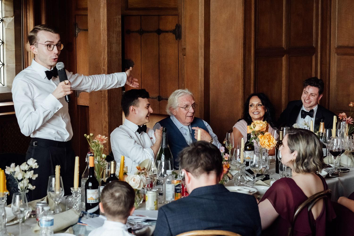 A young man gives a speech at an elegant wedding reception dinner, holding a microphone before seated guests.