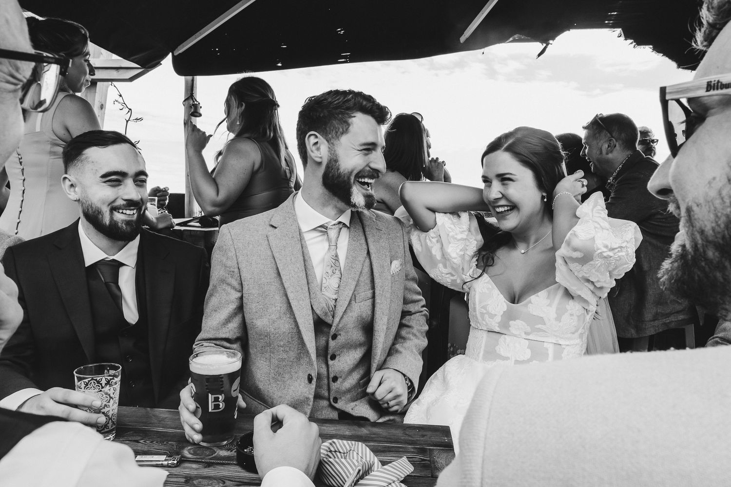 A joyful candid moment at a wedding reception as couple laughs while seated with guests at outdoor venue.