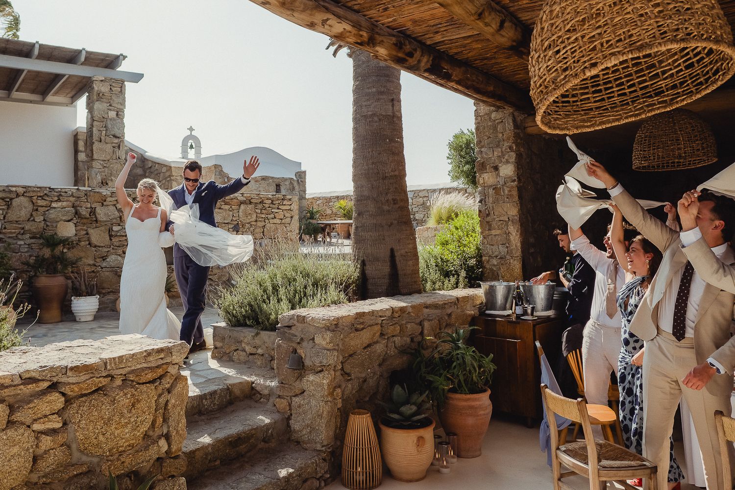Wedding guests gather under woven rattan pendant lights in a rustic Mediterranean courtyard setting during celebration.