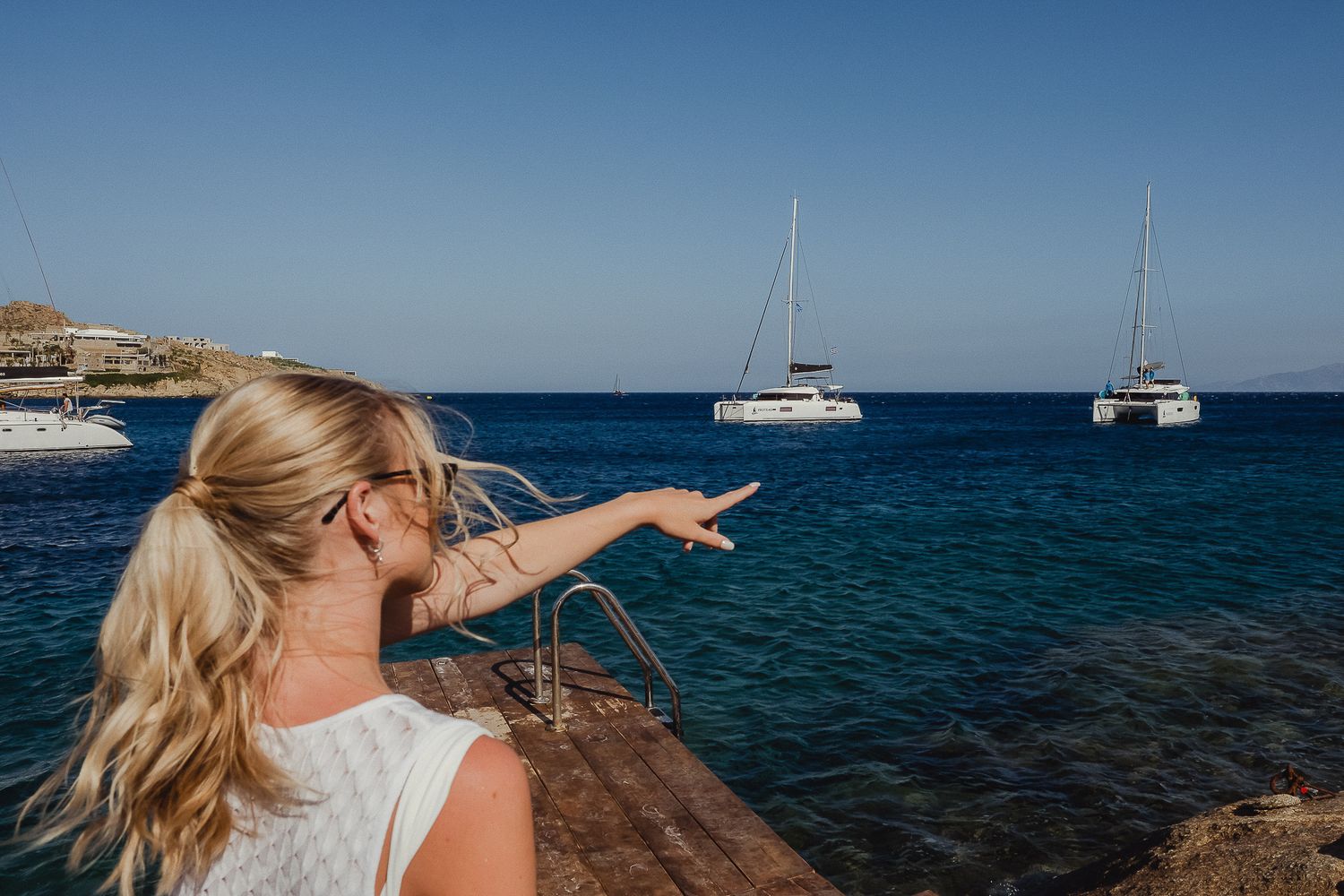 Sailboats anchored in turquoise Mediterranean waters near rocky coastline with clear blue sky above.