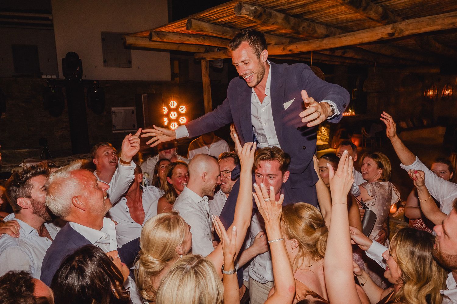 Wedding reception guests dancing and celebrating on a dimly lit dance floor with hands raised in joyful celebration.