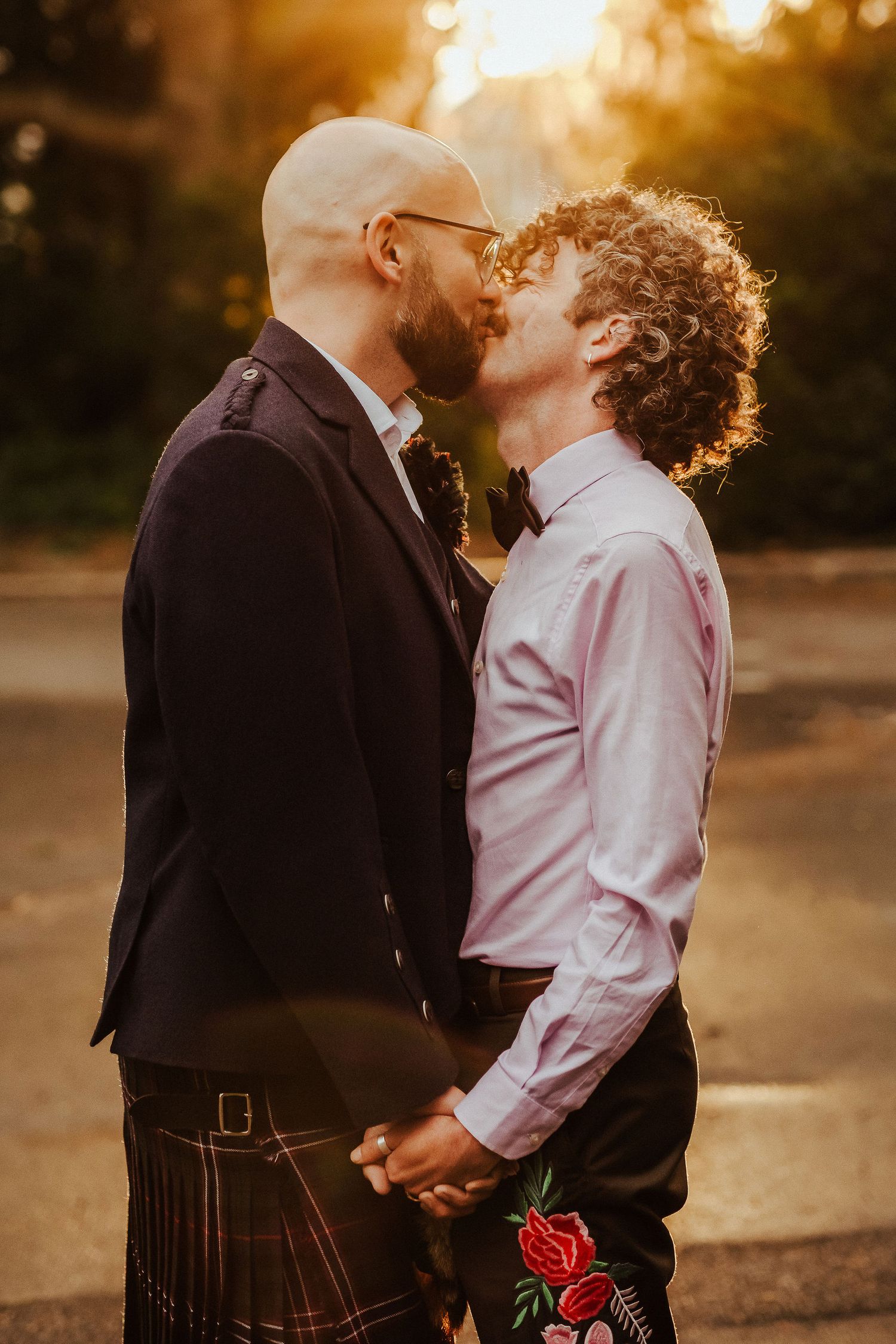 Two grooms in wedding attire, one in a tartan kilt and jacket, share a kiss while holding hands in warm golden sunset light.