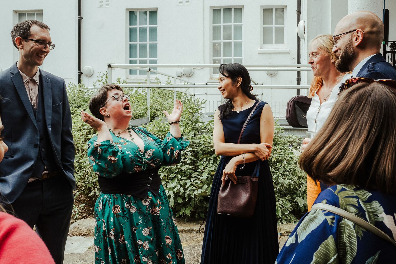 Group of smiling friends chatting and laughing outdoors in a courtyard, with a woman in a green floral dress gesturing animatedly.