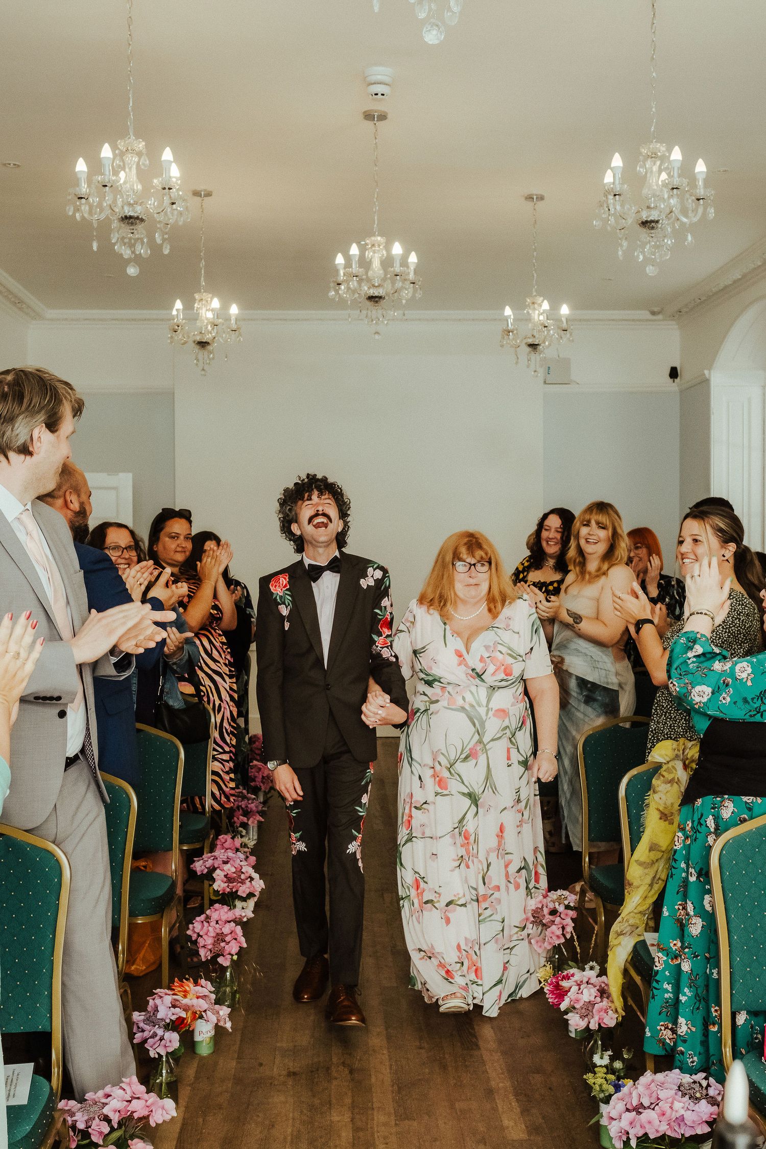 Smiling newlyweds walk hand in hand down a flower-lined aisle as cheering wedding guests clap beneath crystal chandeliers.