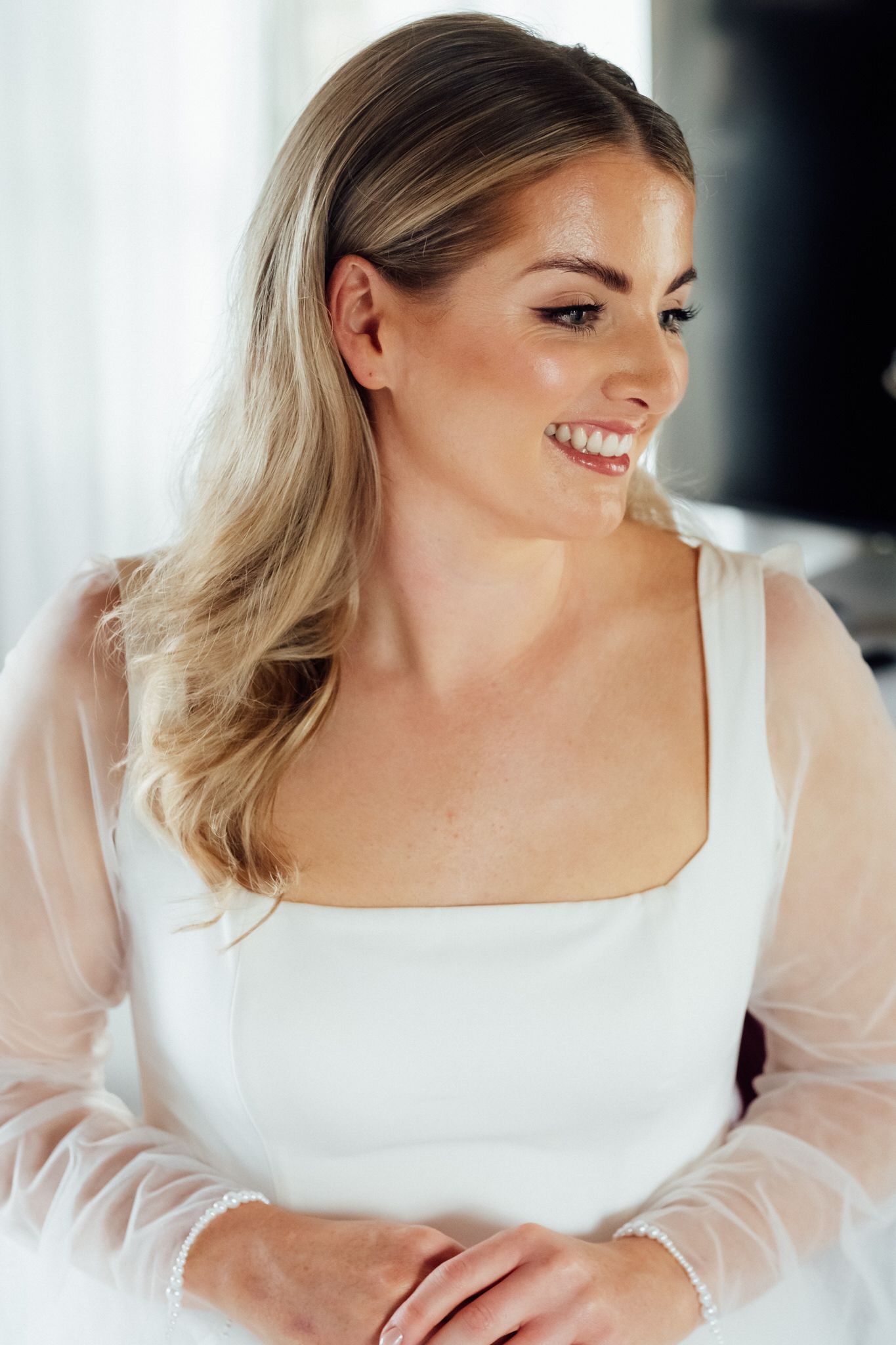 Smiling bride with long wavy blonde hair wears a white square-neck wedding gown with sheer tulle sleeves, glancing aside with hands folded.